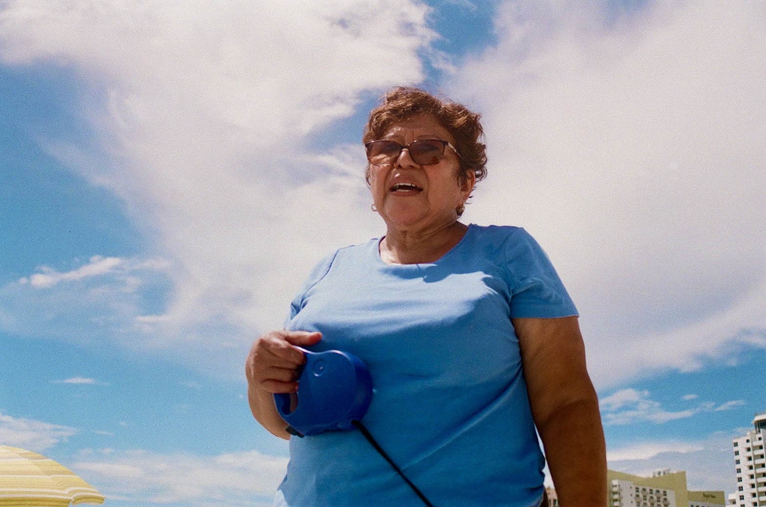Older woman with short curly hair wearing sunglasses and a blue shirt, holding a blue object, outdoors under a partly cloudy sky.