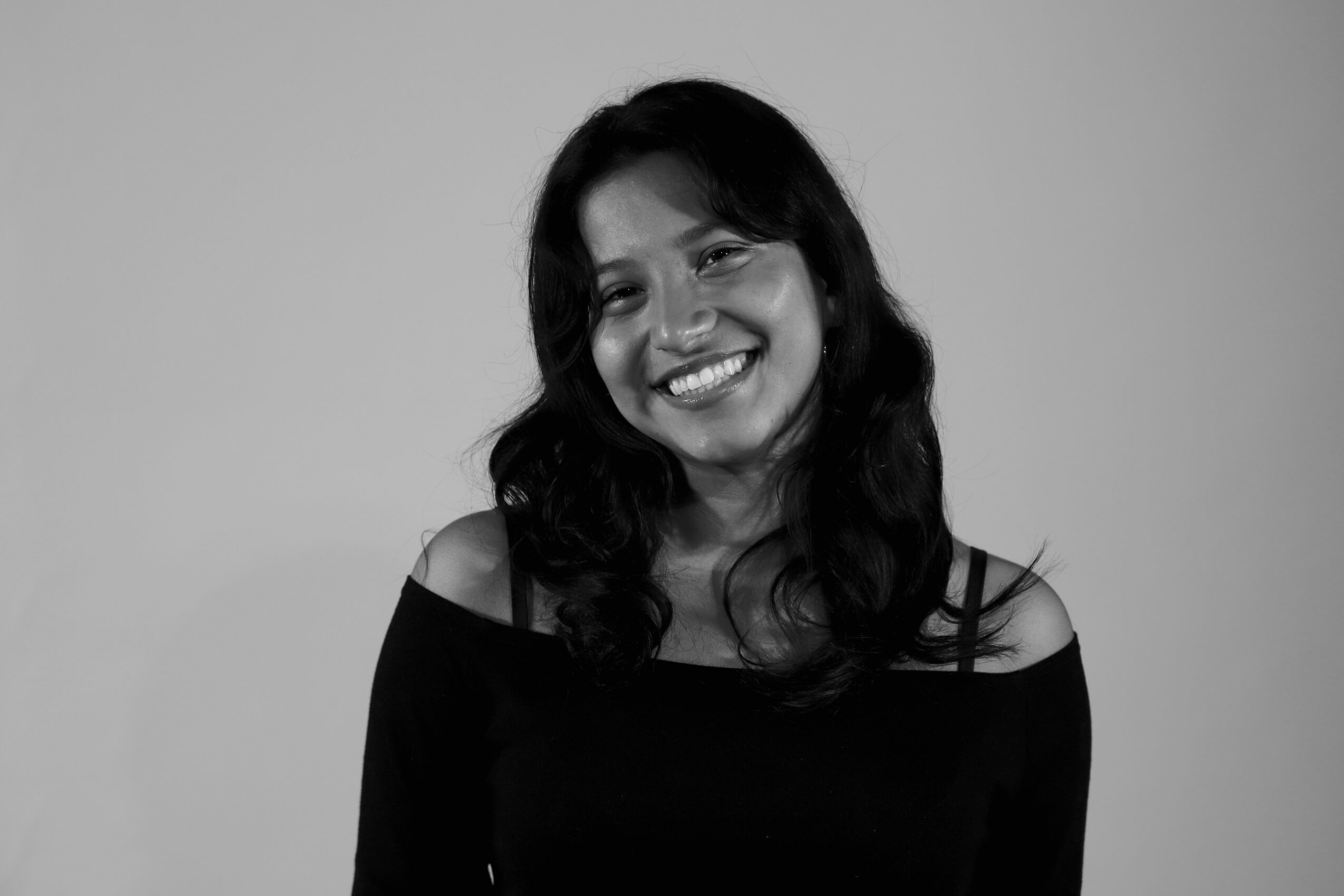 Smiling woman with dark hair wearing a black off-shoulder top, facing forward, in grayscale.