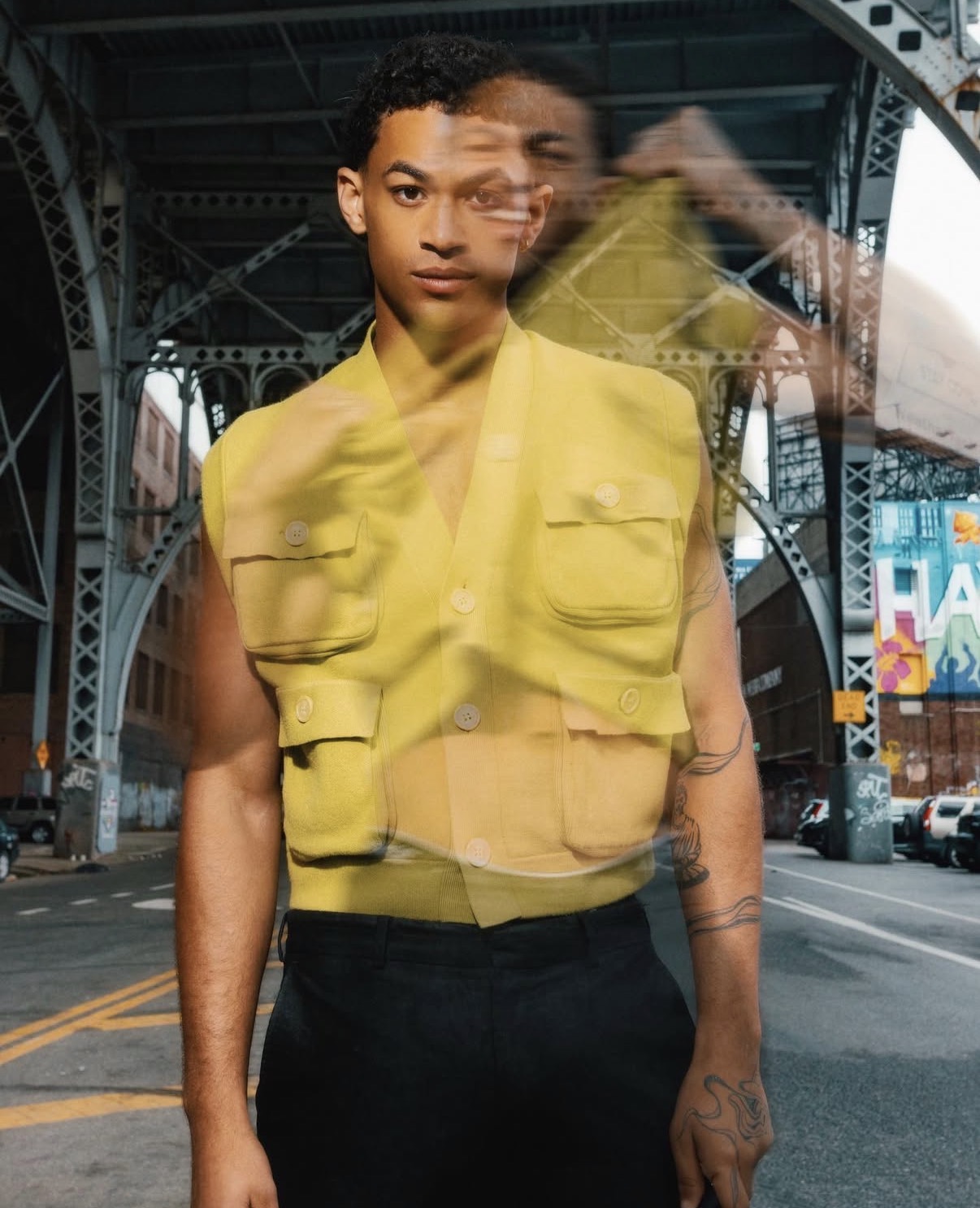Person standing under a bridge wearing a yellow vest, with a city street background, multiple reflections visible.