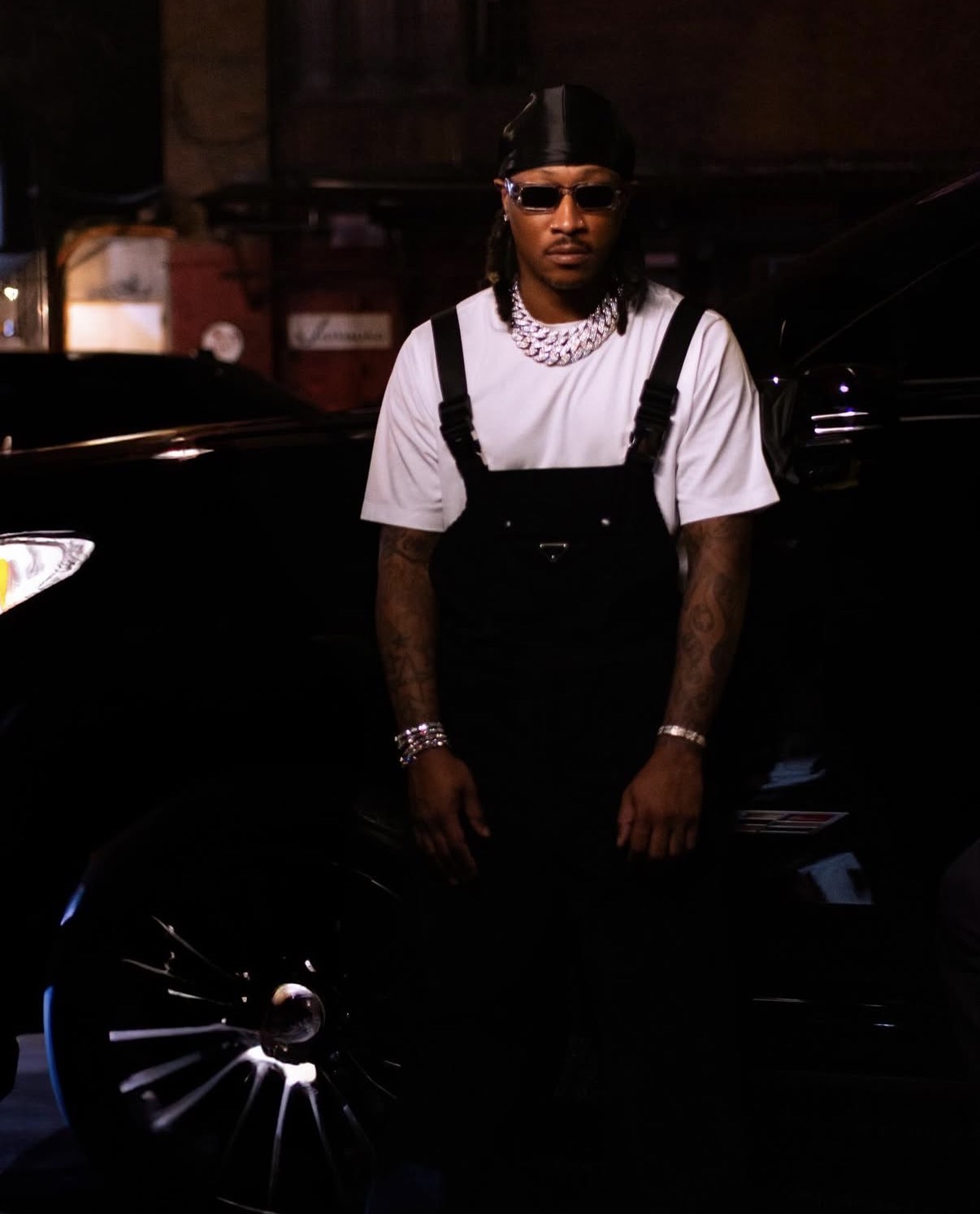 Person wearing sunglasses, a white t-shirt, black overalls, and jewelry, standing in front of a black car at night.