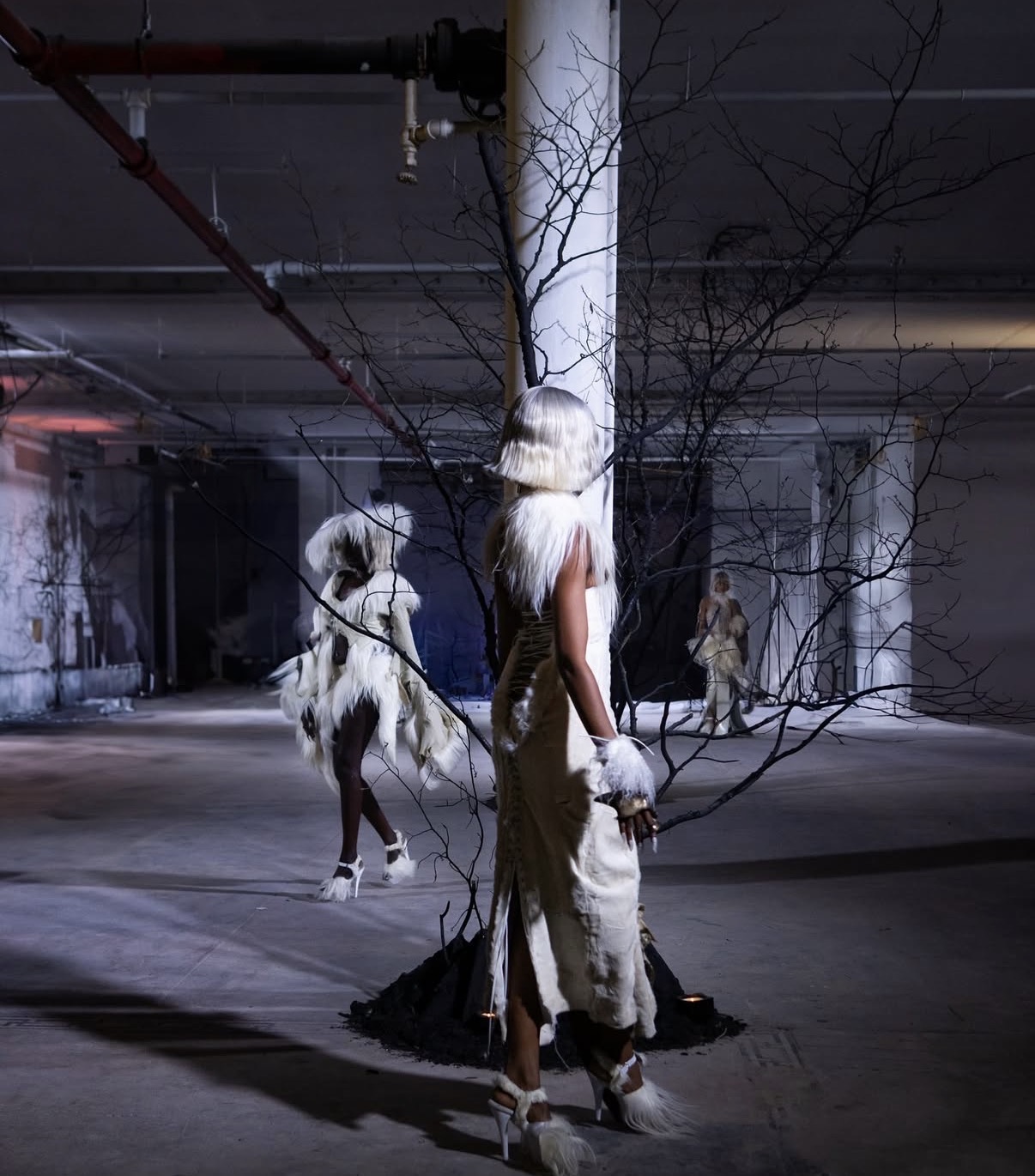 Person in a tall, white feathered headdress and long dress standing in an underground parking garage with a horse nearby.