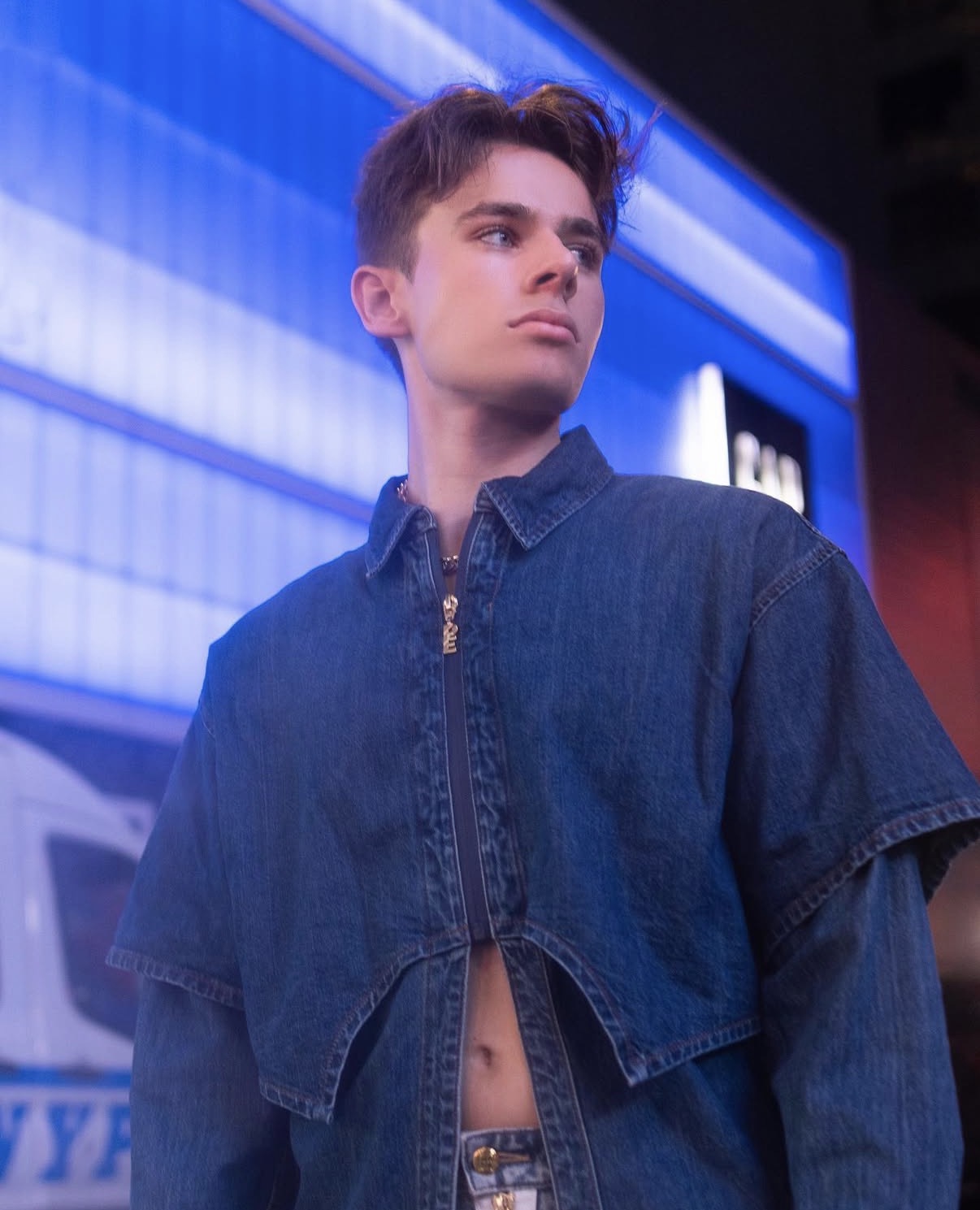 Young man with short hair wearing a denim jacket, standing outdoors at night with illuminated buildings behind him.