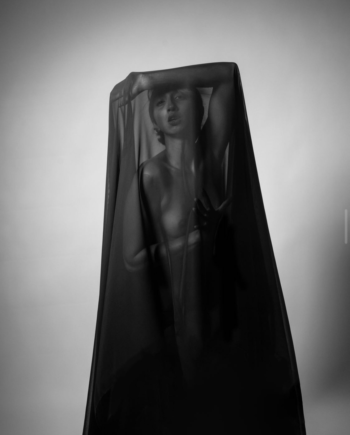 Person behind a sheer black fabric, with arm raised and face partially visible, in a studio setting.
