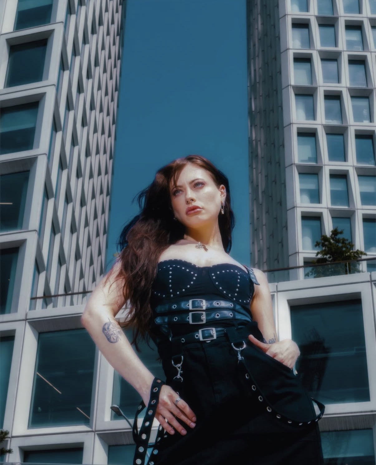 Woman with long hair standing between tall modern buildings, looking at the camera.