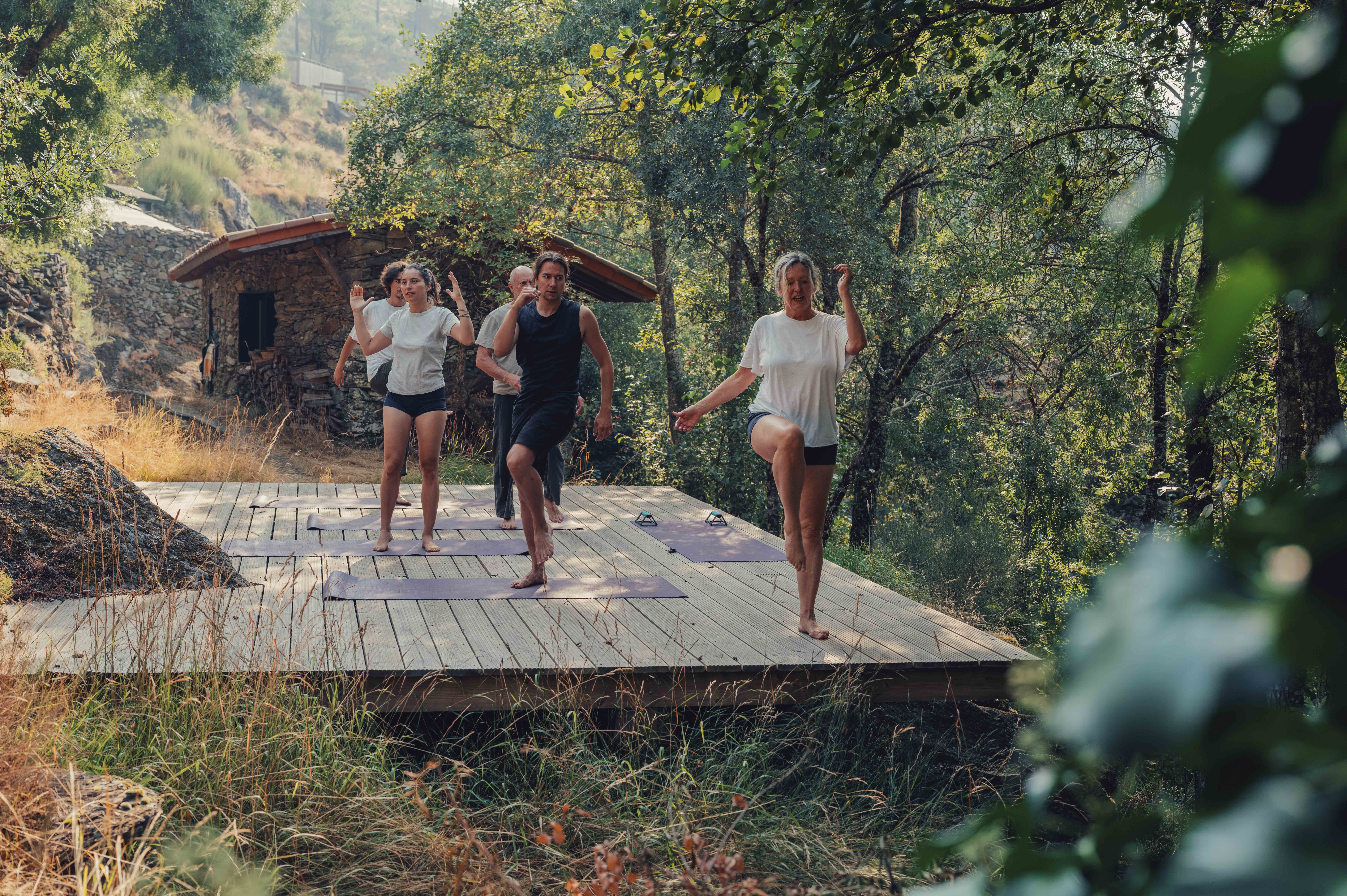 Four people run on a wooden platform outdoors with trees and a small building in the background.