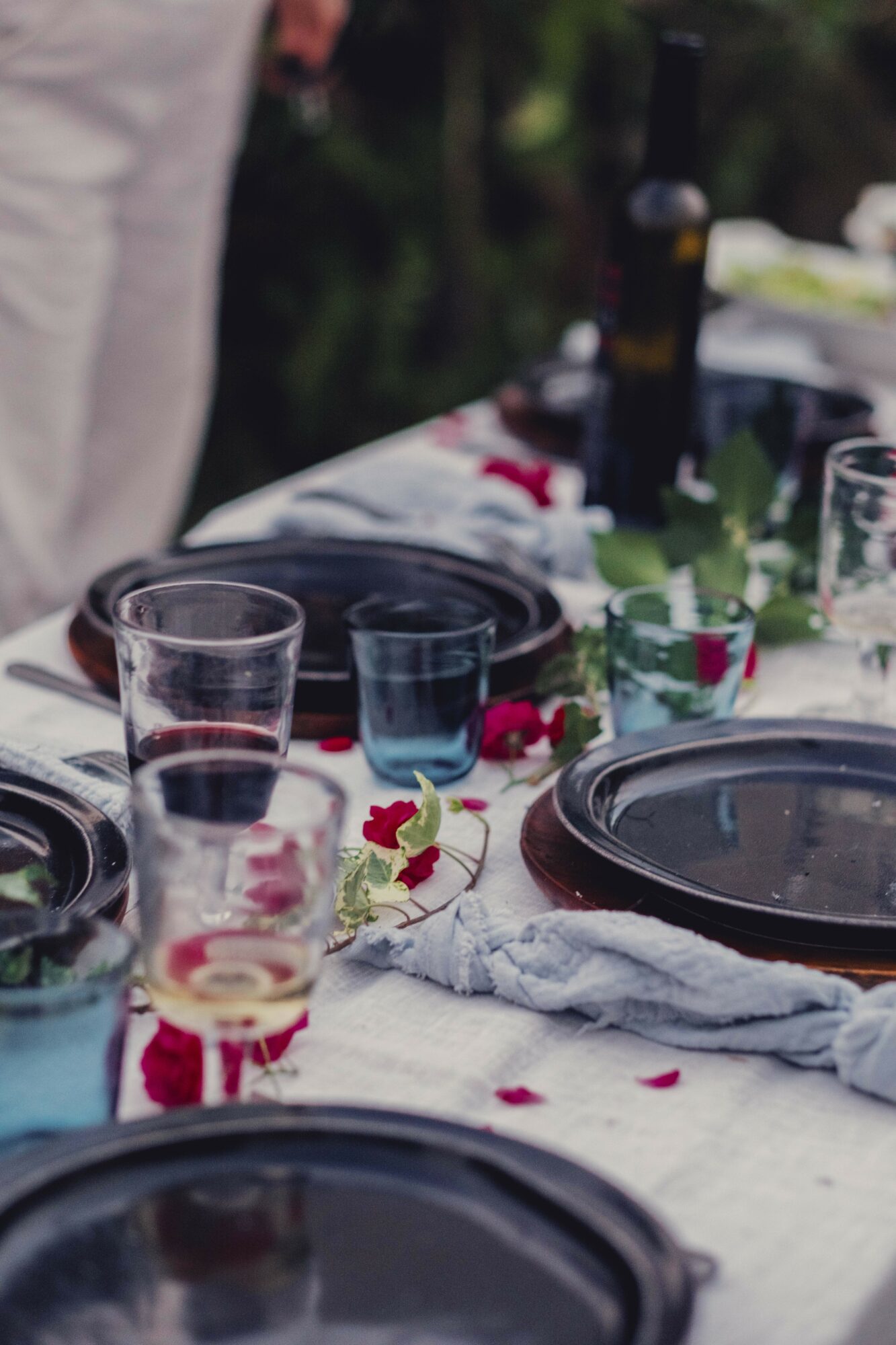 Table set with black plates, glasses, a bottle, and scattered flower petals outdoors, with greenery in the background.