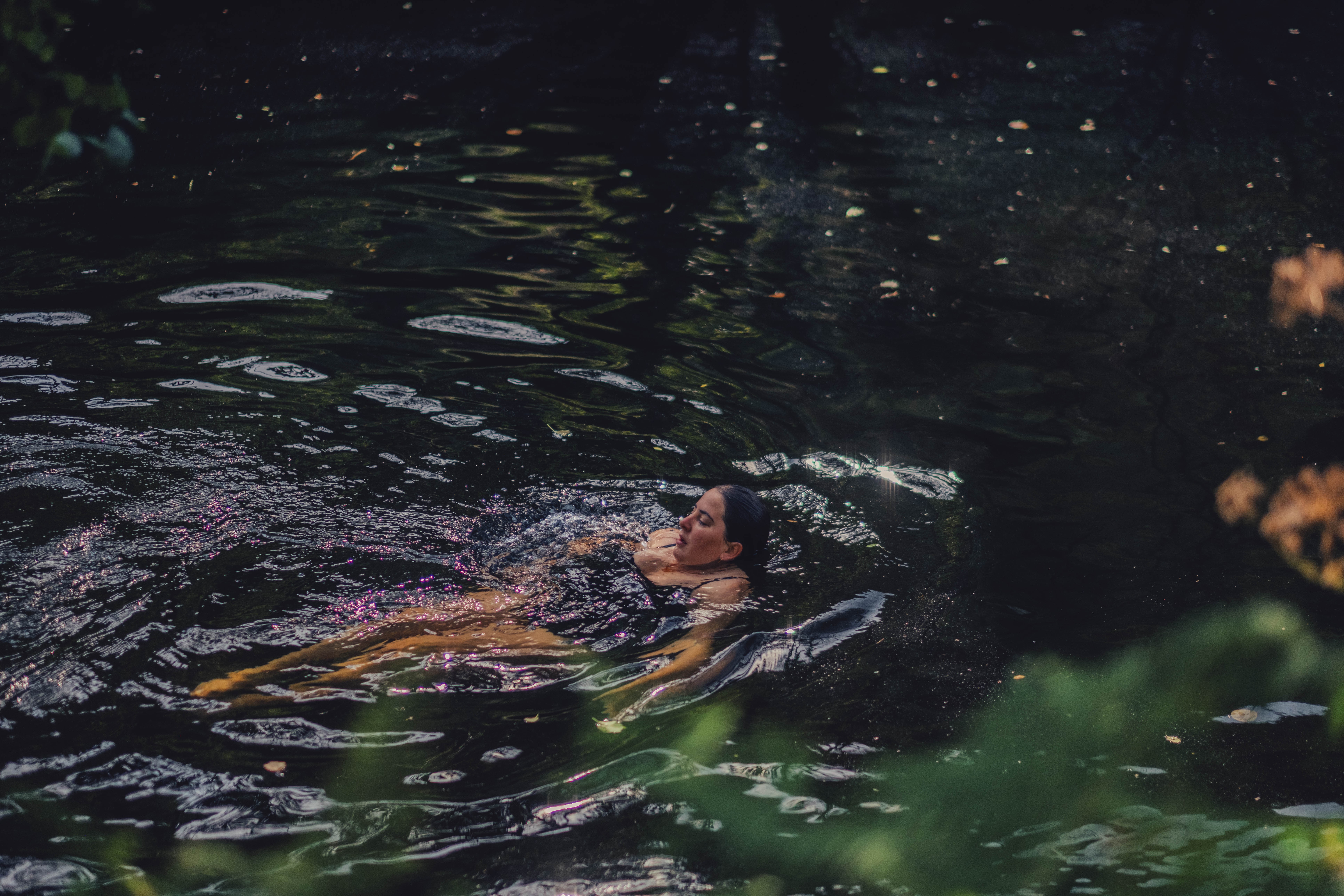 Person floating on water with head above surface, surrounded by dark water and some greenery.