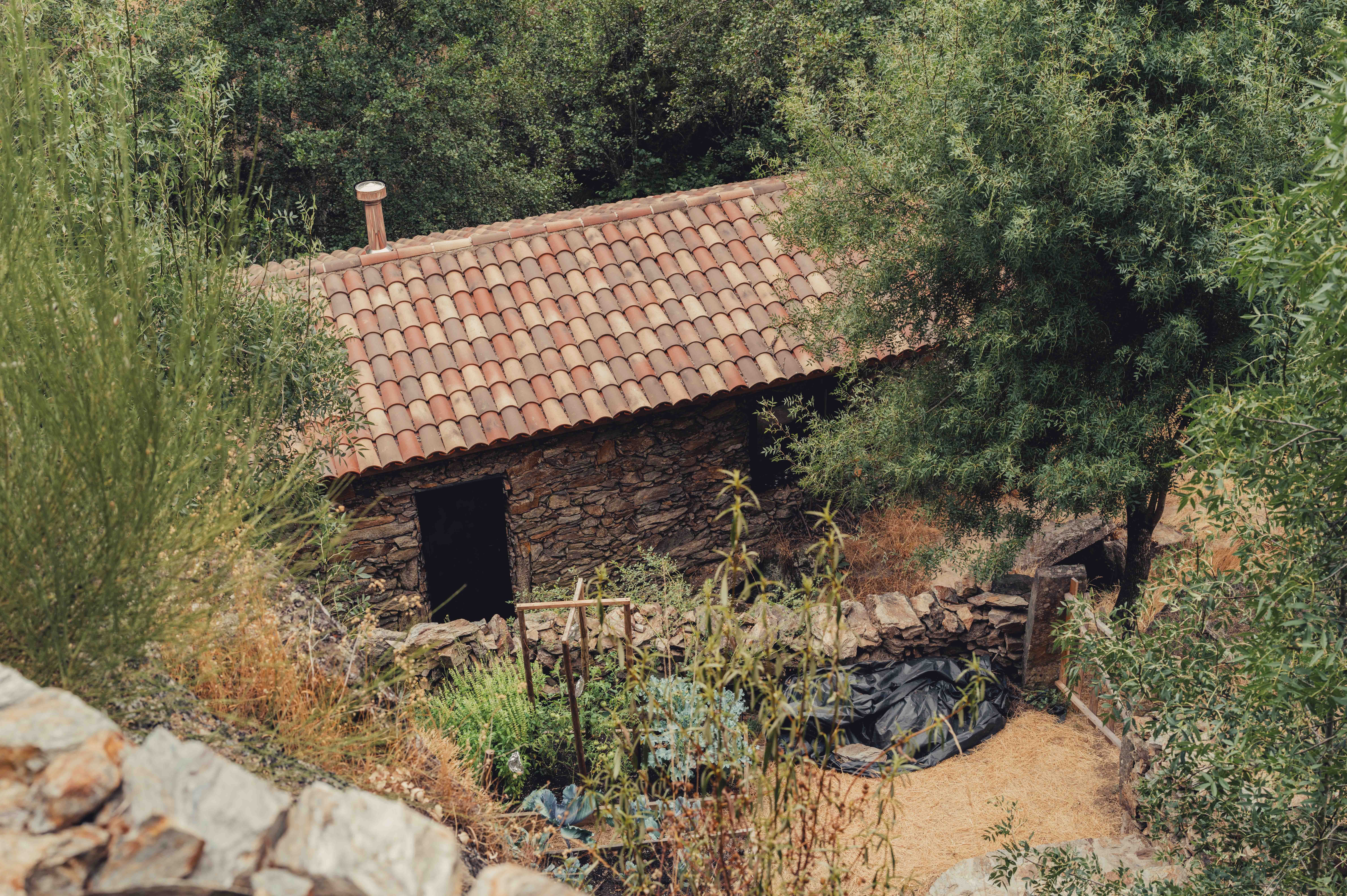 A small stone building with a tiled roof is surrounded by trees and vegetation, with a garden area in front.