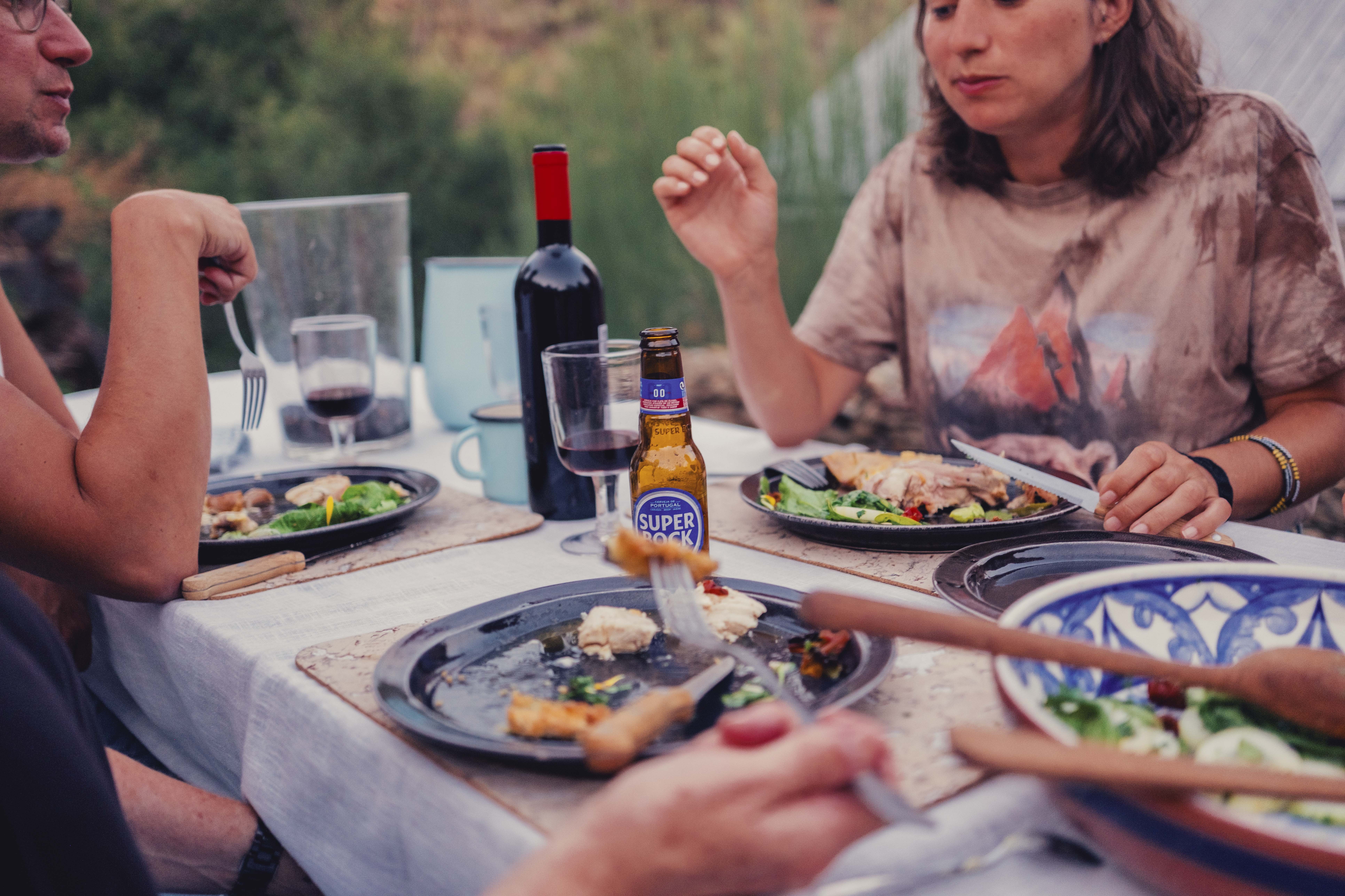 People dining outdoors with plates of food, wine, and drinks on a table, with a blurred natural background.