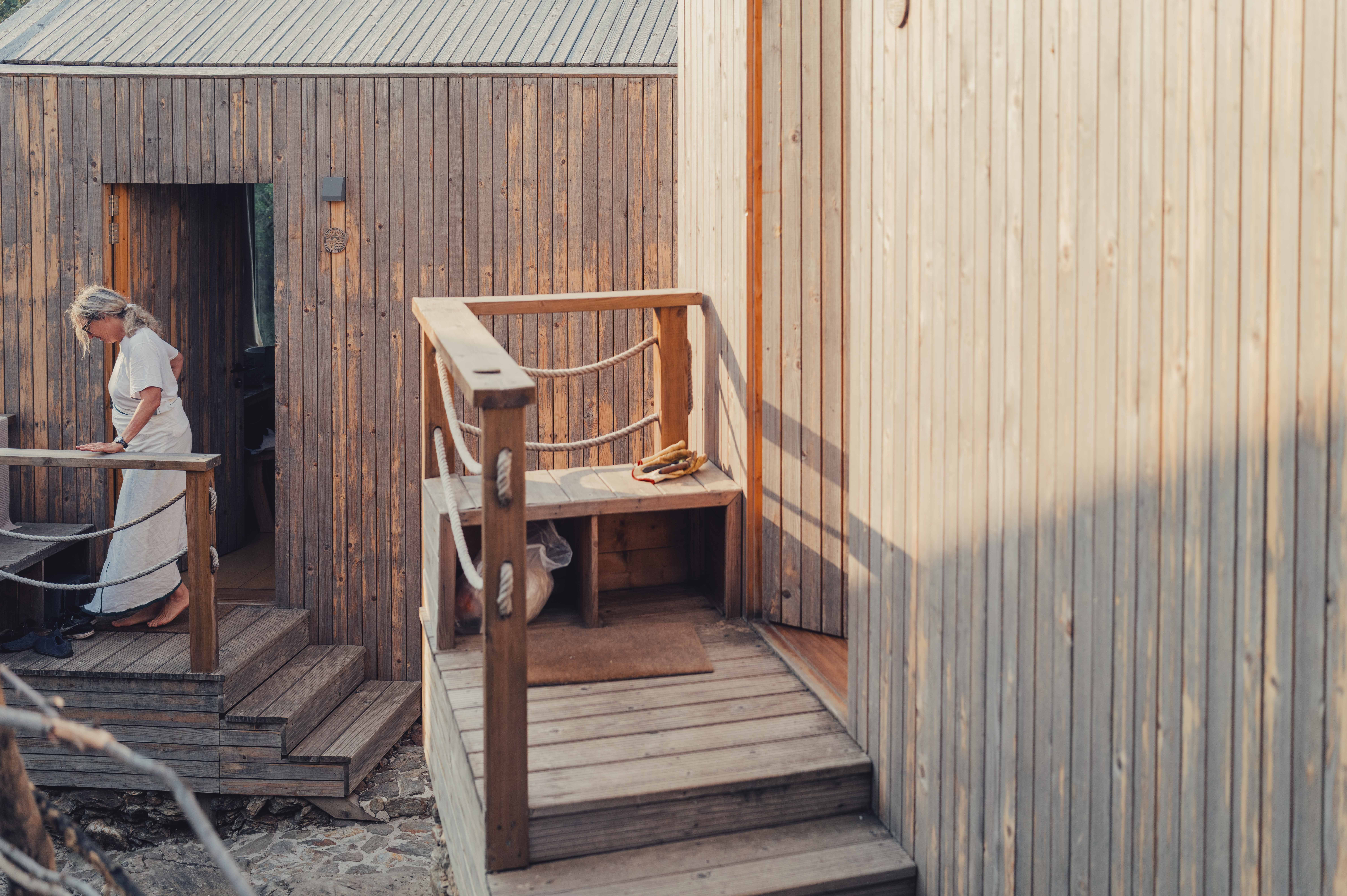 Wooden stairs leading to a small platform outside a building, with a person in the background near a door, sunlight casting shadows.