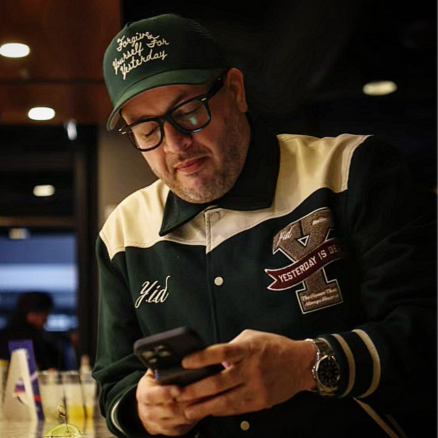 Man wearing glasses, cap, and jacket looking at a smartphone in a dimly lit indoor setting.