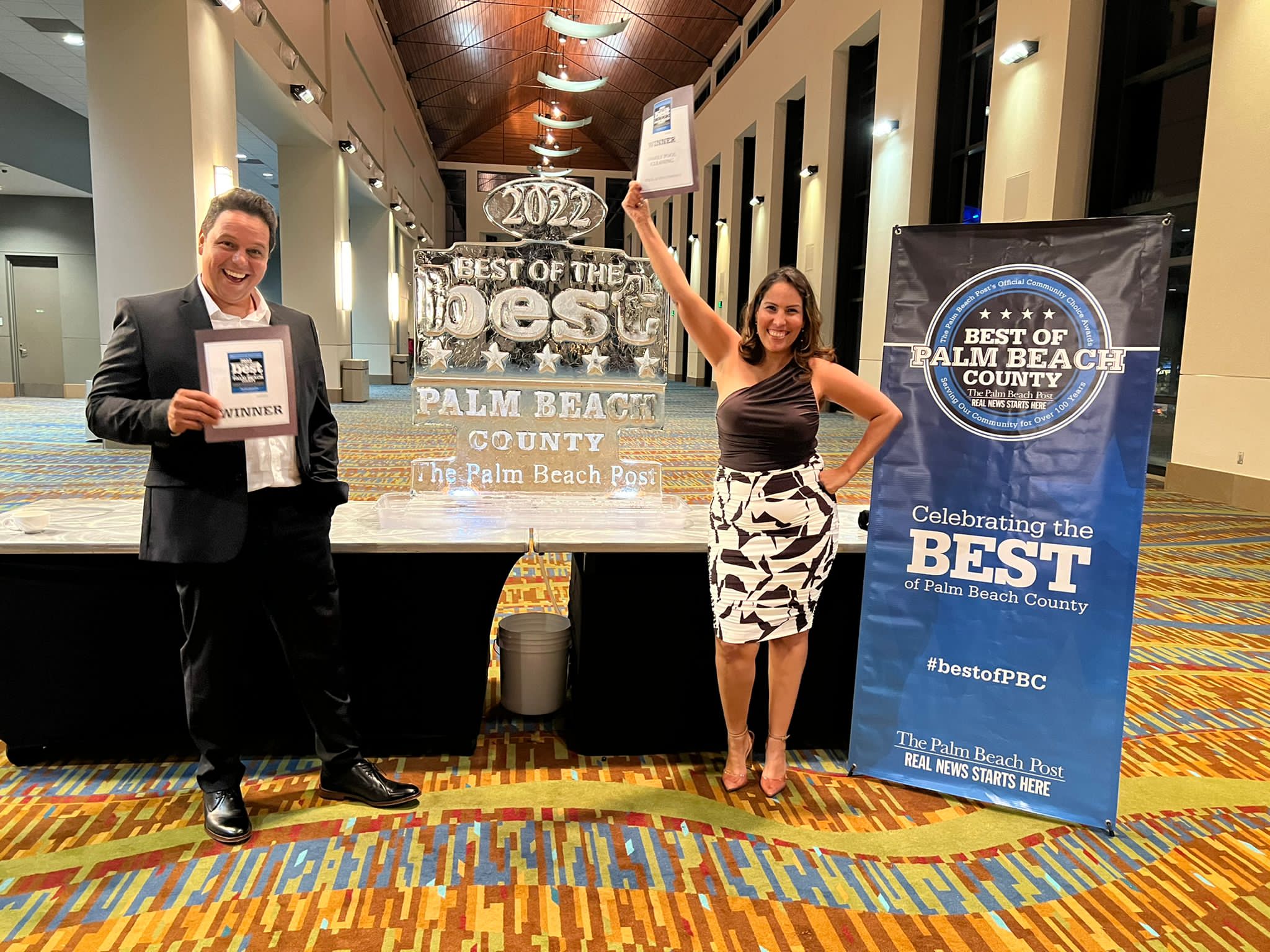 Two people standing indoors next to a table with awards and a banner, smiling at the camera.