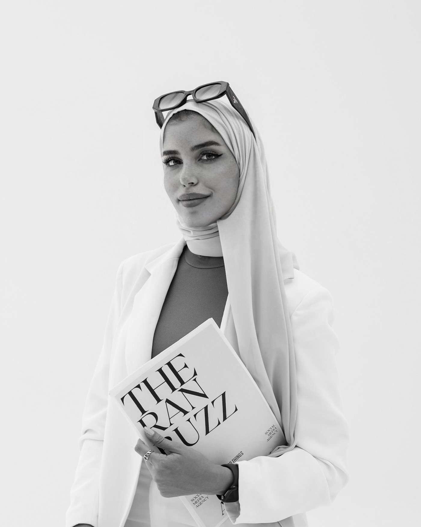Woman wearing hijab and glasses holding a magazine titled 'The Rank Buzz' against a plain background.