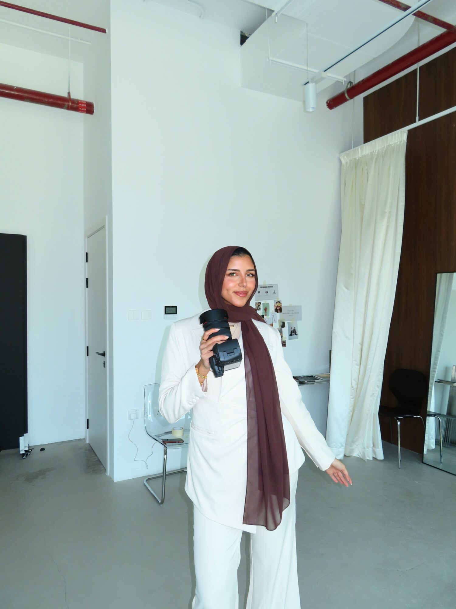 Woman with a camera taking a selfie in a bright room with white walls and a curtain.