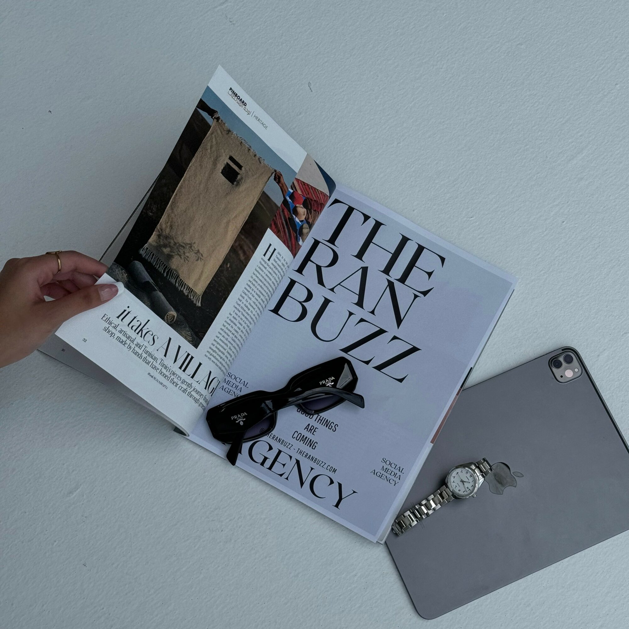 Open magazine with a black strap, a silver watch, and a gray smartphone on a white surface.