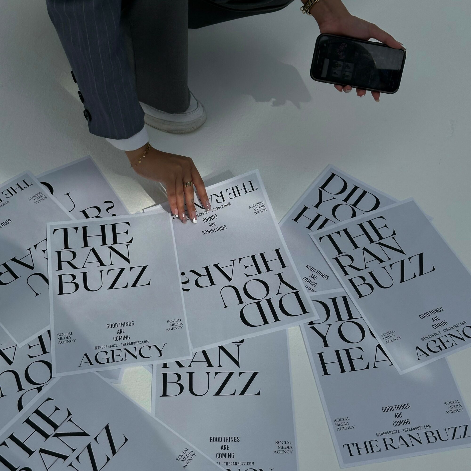 Person arranging newspapers on a table, holding a phone in one hand, newspapers titled 'THE RAN BUZZ' and 'THE RAN BUZZ'.