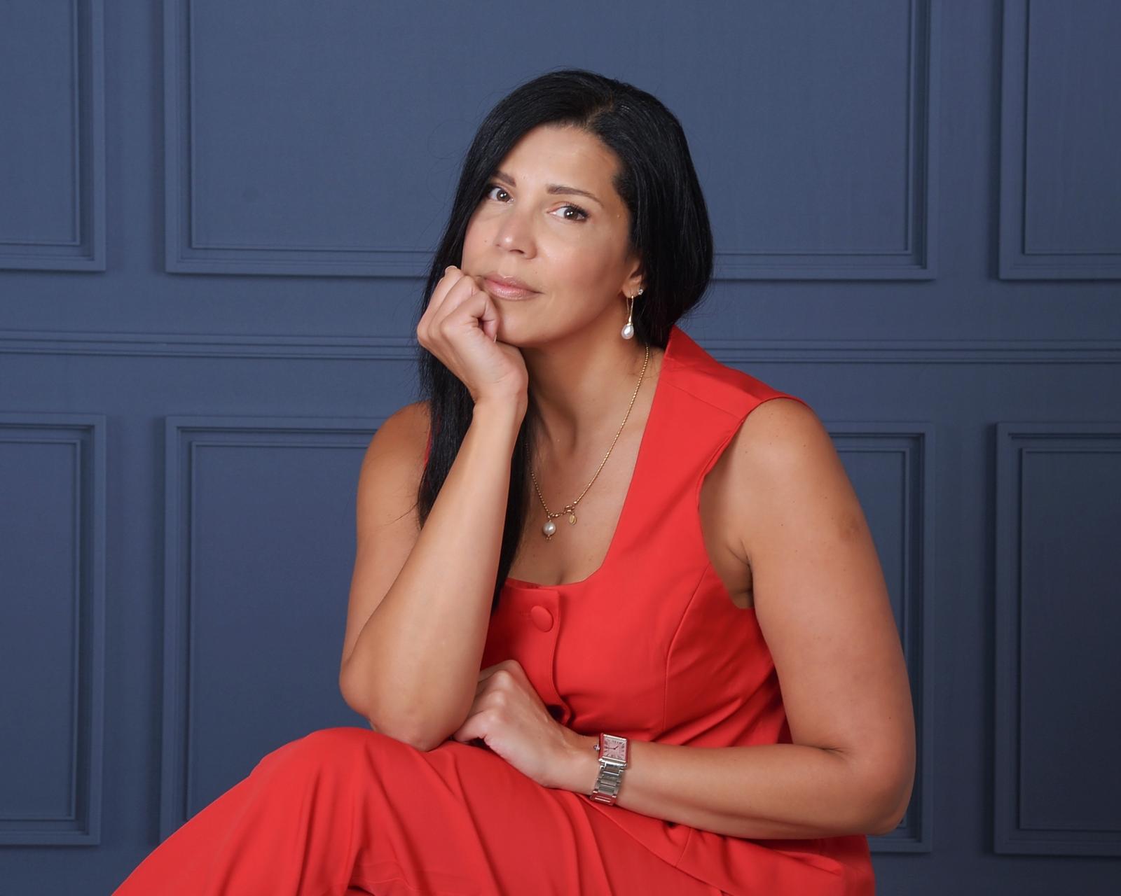 Woman with long dark hair in a red dress, sitting with her hand on her chin, against a blue paneled wall.