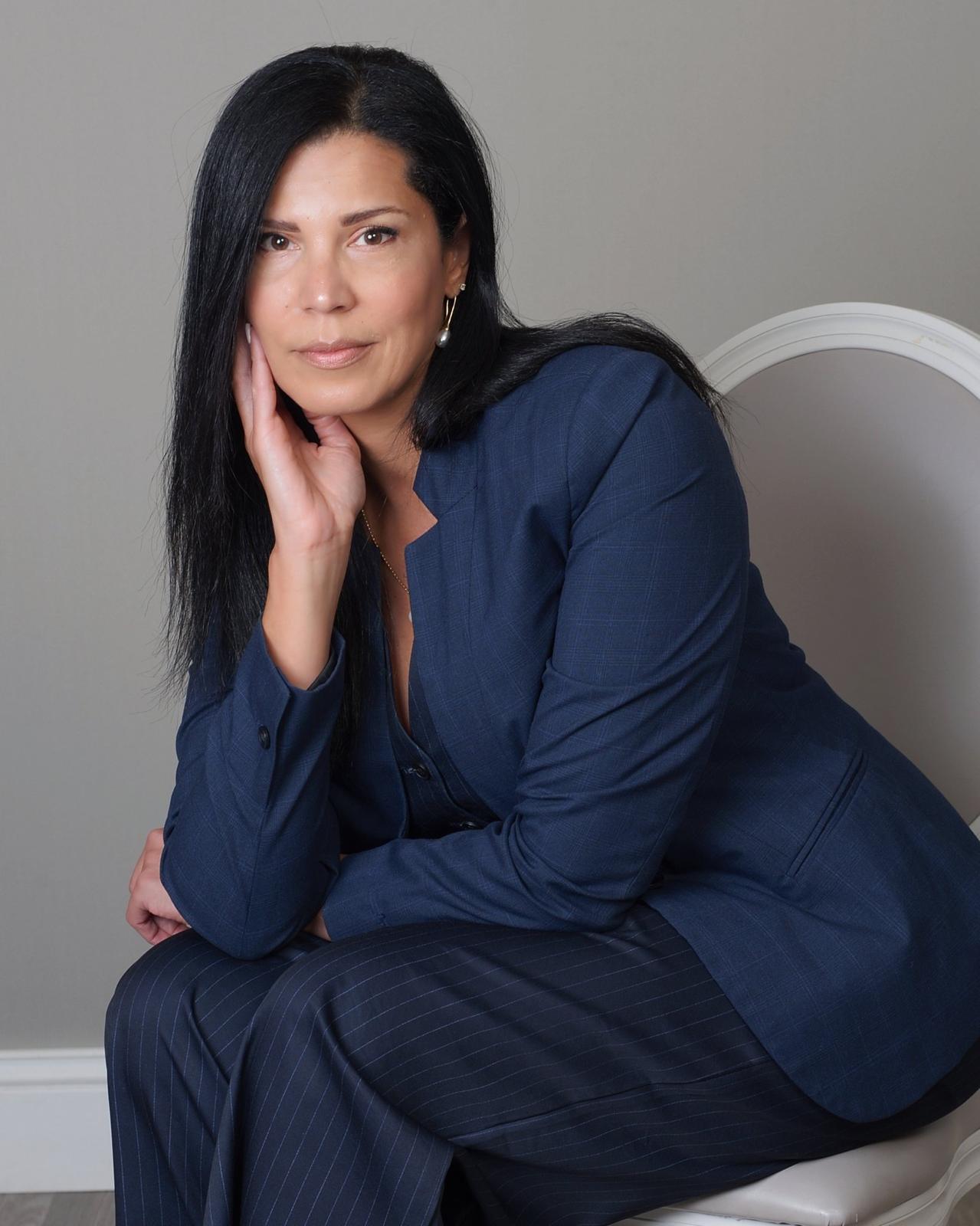 Woman with long dark hair in a blue blazer and pants, sitting and resting her chin on her hand.