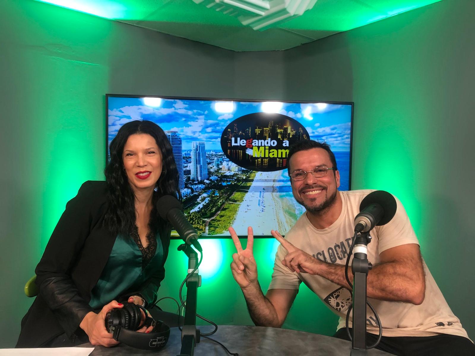 Two people sit at a table with microphones, smiling, in a green-lit studio with a cityscape background on a screen.
