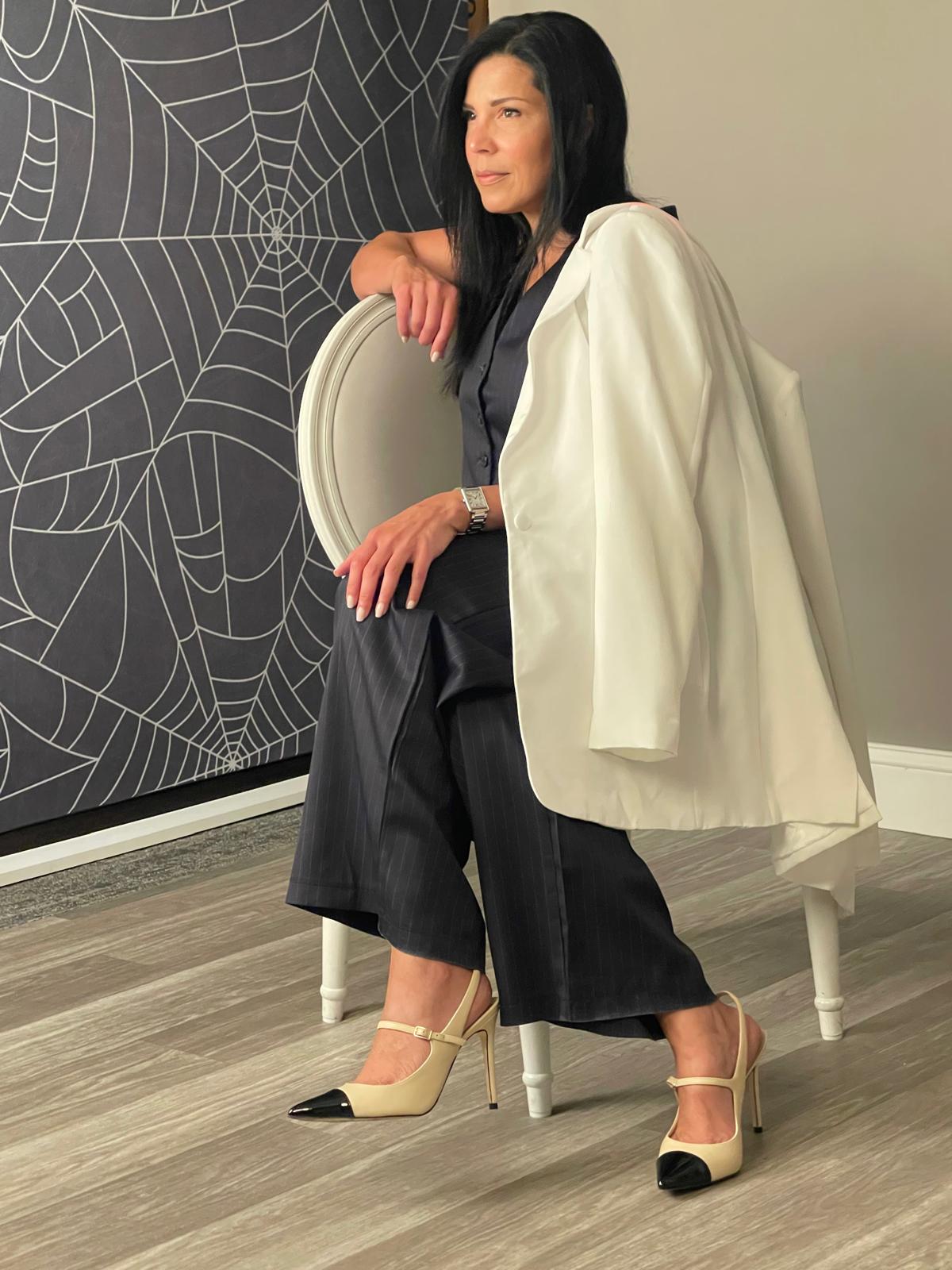 Woman with black hair in black and white outfit, sitting on a white chair, in front of a spider web artwork.