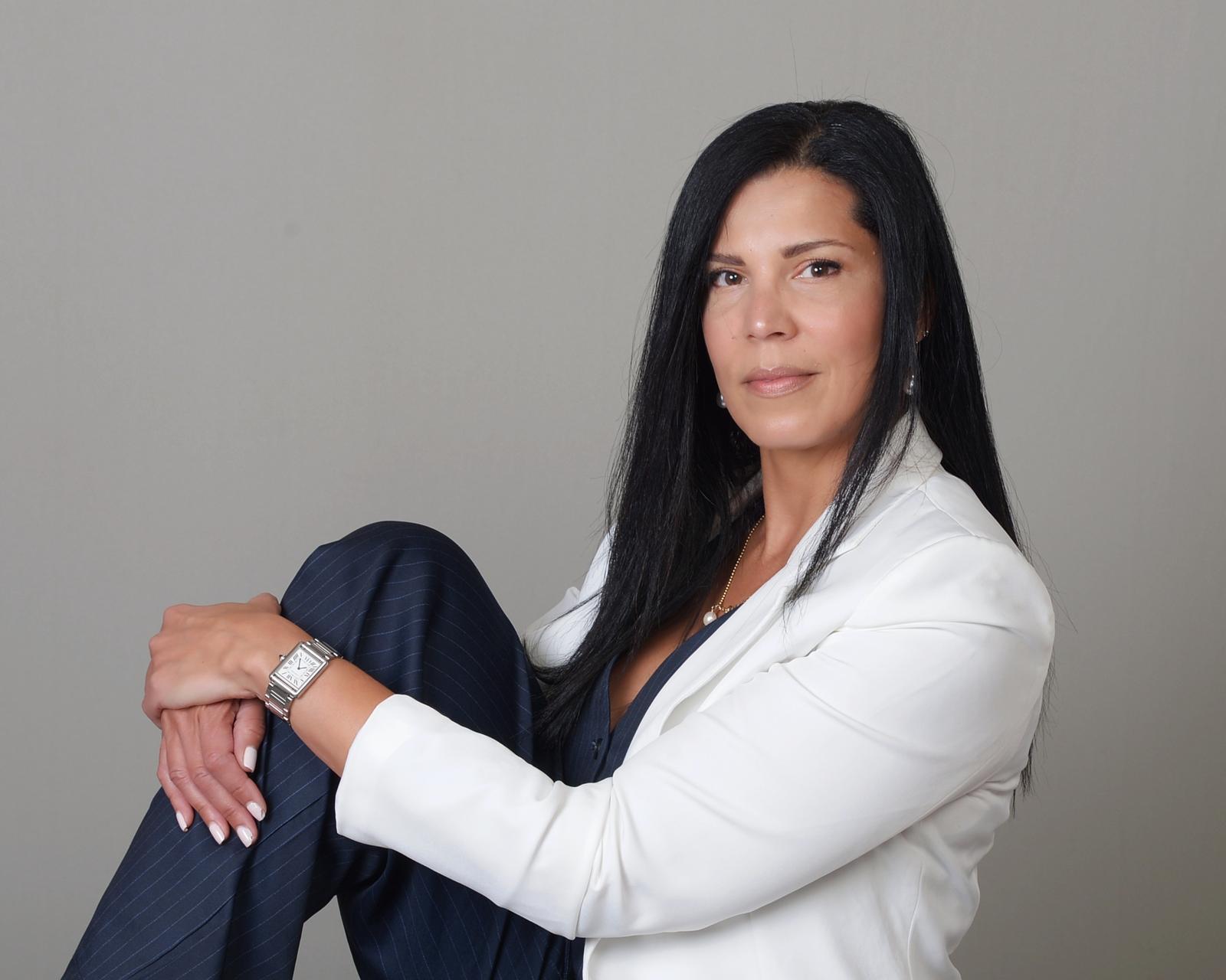 Woman with long black hair wearing a white blazer and dark pants, sitting against a plain gray background.