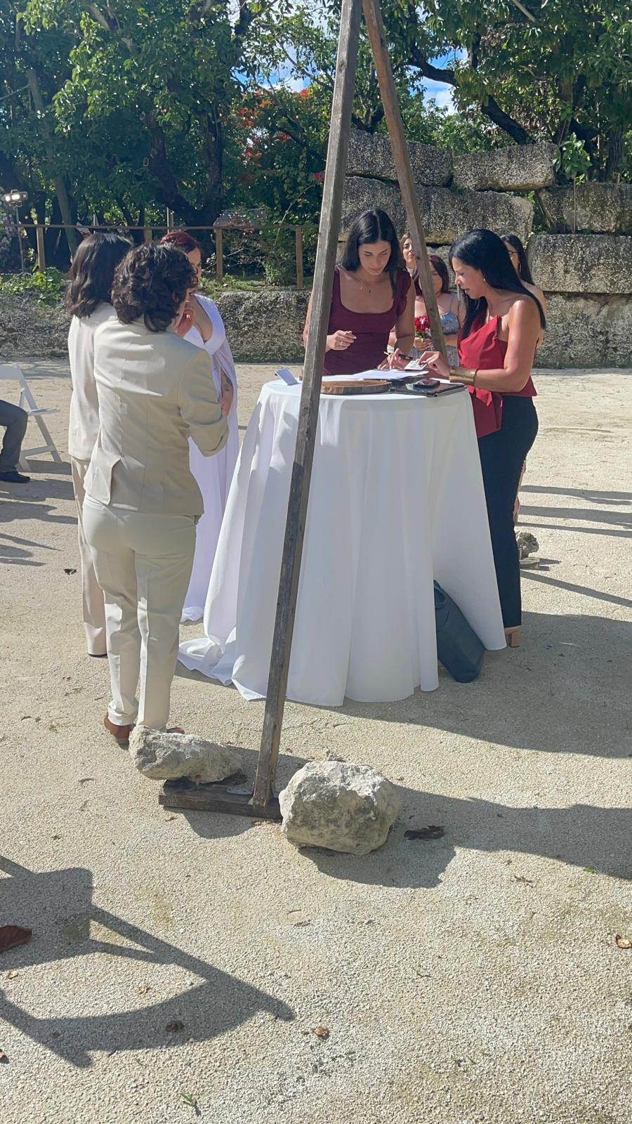 Four women gather around a table outdoors, with trees and a fence in the background.