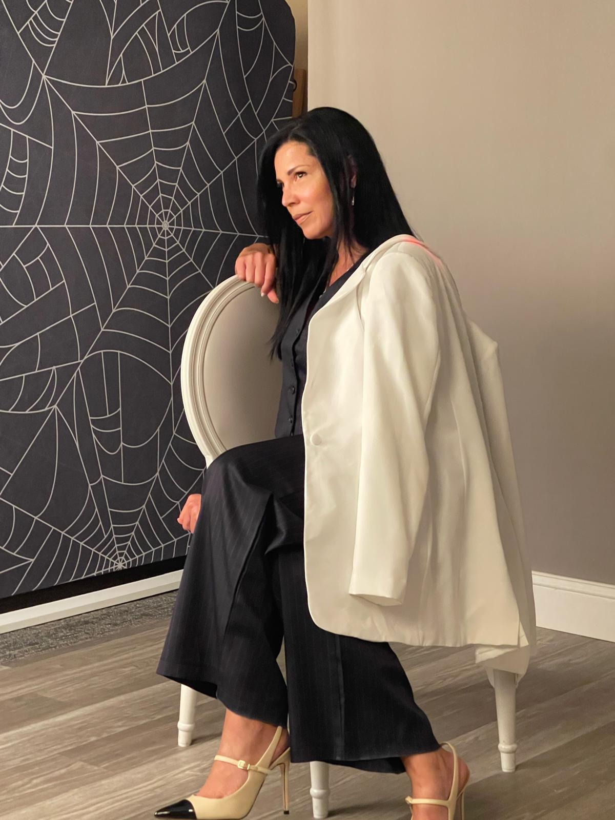 Woman sitting on a white chair, wearing a black outfit and a white jacket, with a spider web artwork behind her.