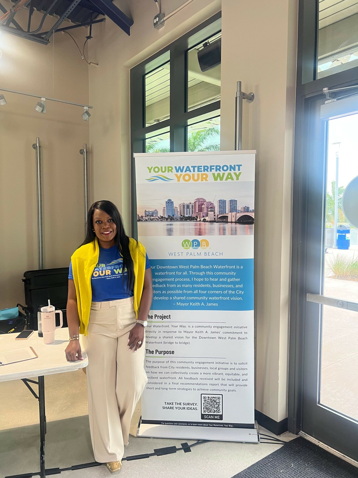 Woman standing next to a water-themed informational poster in a room with large windows and a door, wearing a yellow jacket and blue shirt.