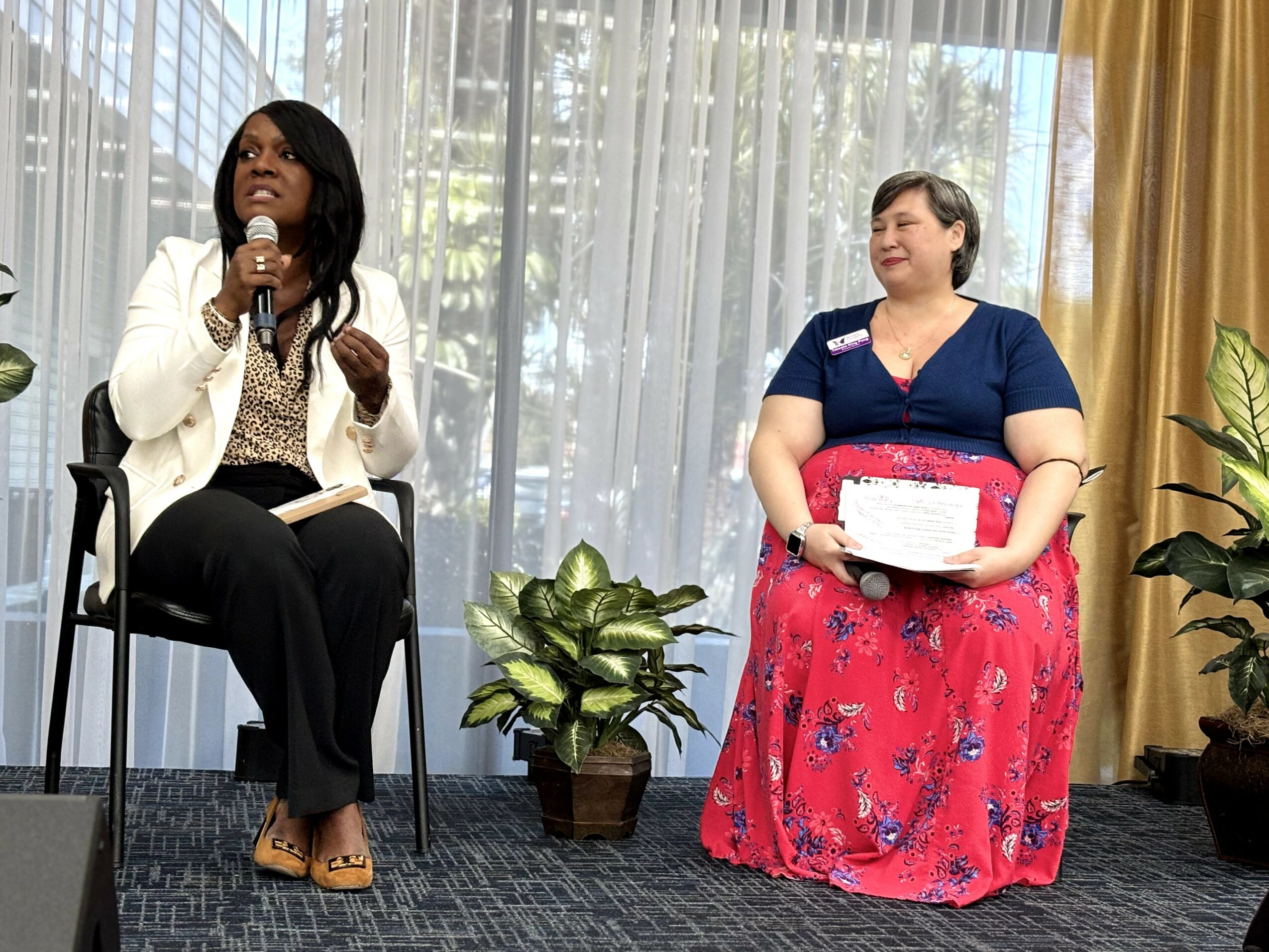 Two women sitting on chairs indoors, one speaking into a microphone, the other holding a box, with plants and curtains behind.