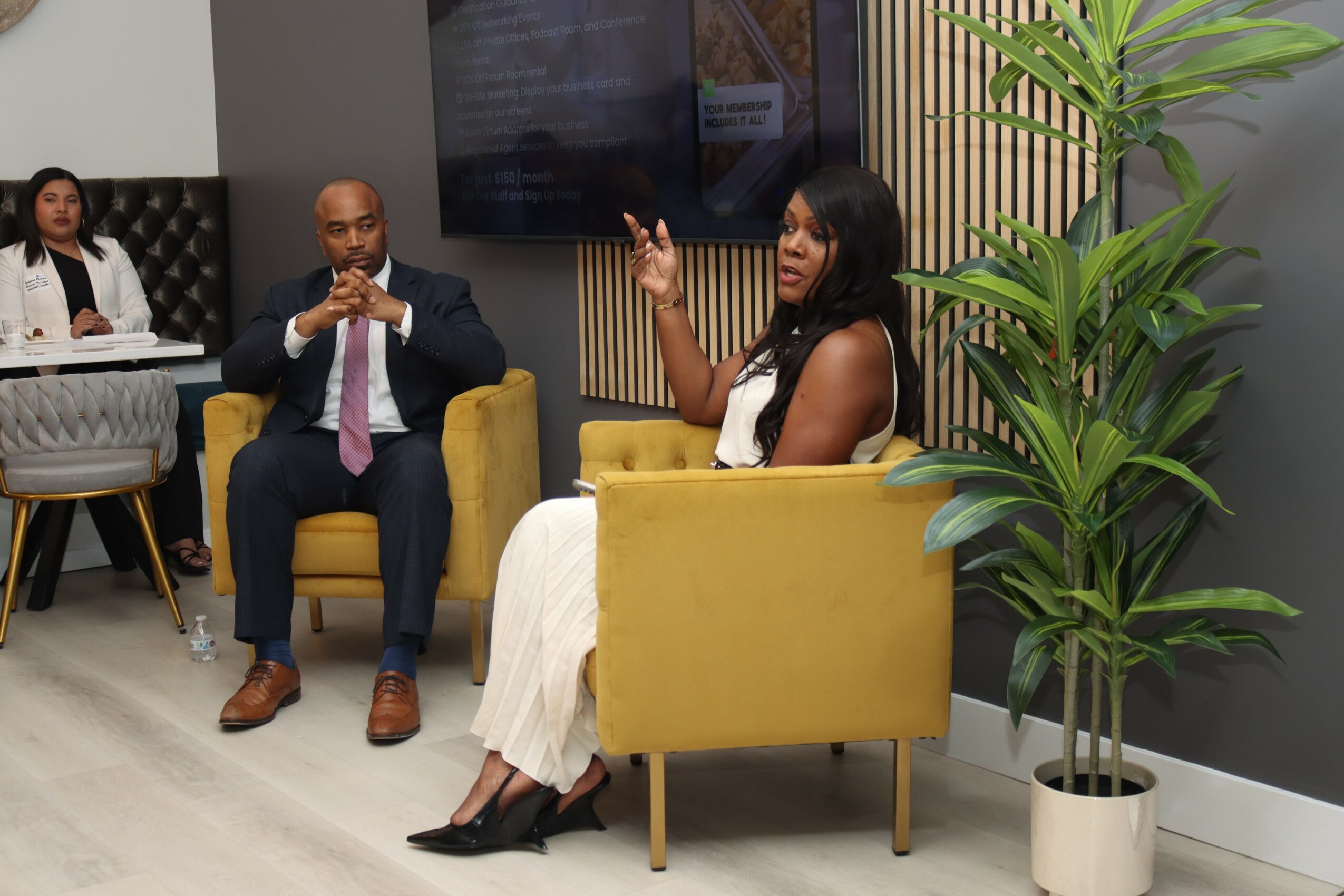 Three people in a modern room, one woman sitting in a yellow chair gesturing, two others seated nearby, and a large plant.