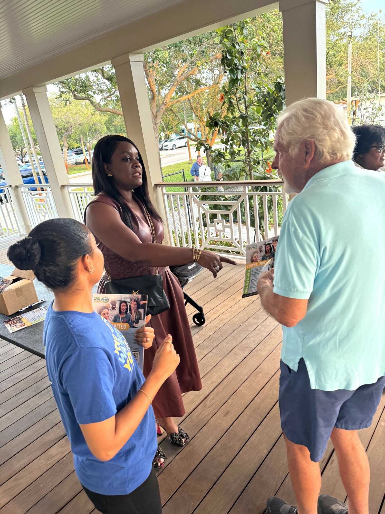 Three people engaged in conversation on a porch with trees and a park in the background.