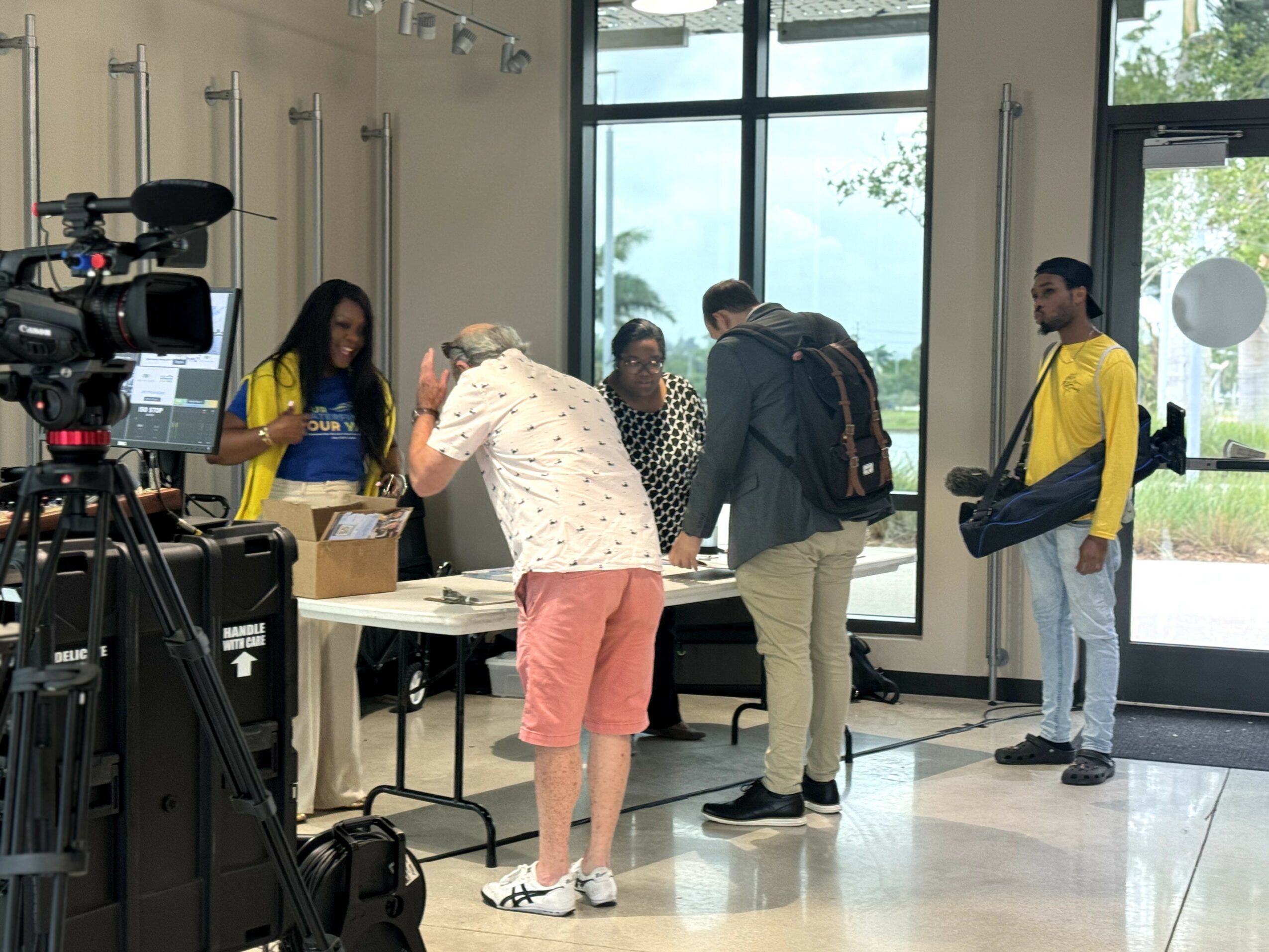 People standing in line at a registration table inside a room with large windows, some with backpacks, and a camera on a tripod.