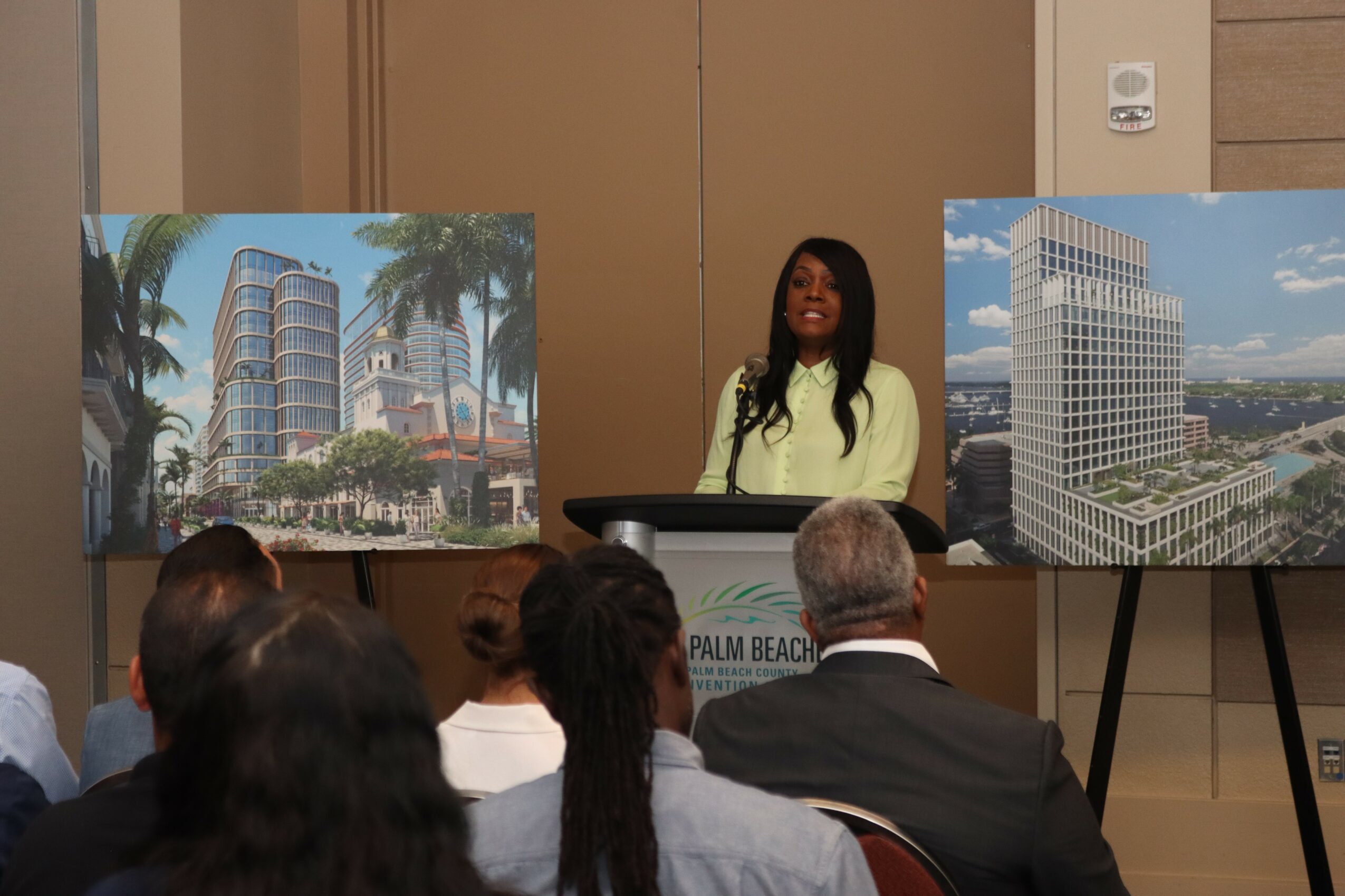 Woman standing behind a podium at a presentation, with two large building images on display, audience seated facing her.