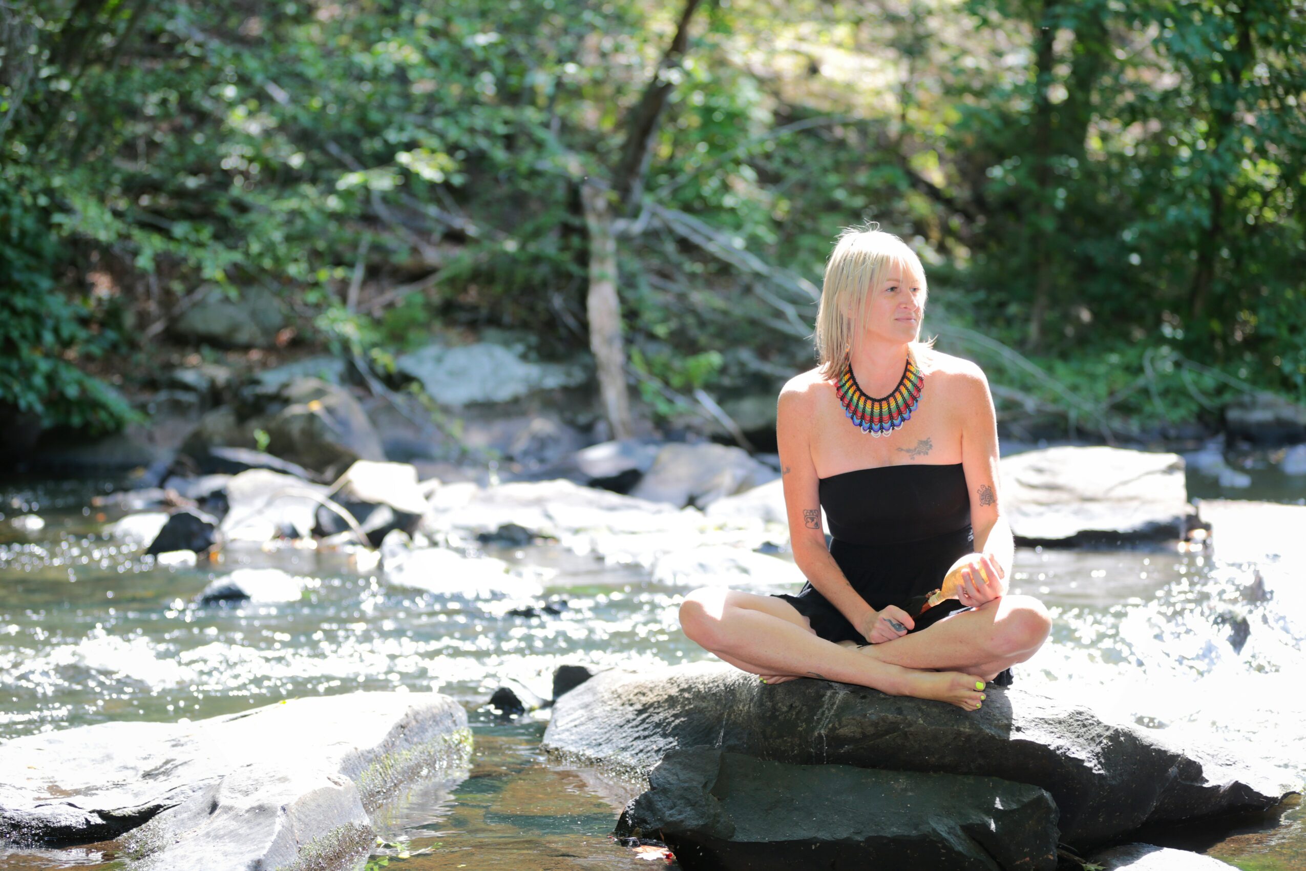 Woman with blonde hair sitting cross-legged on a rock in a river surrounded by trees and rocks.