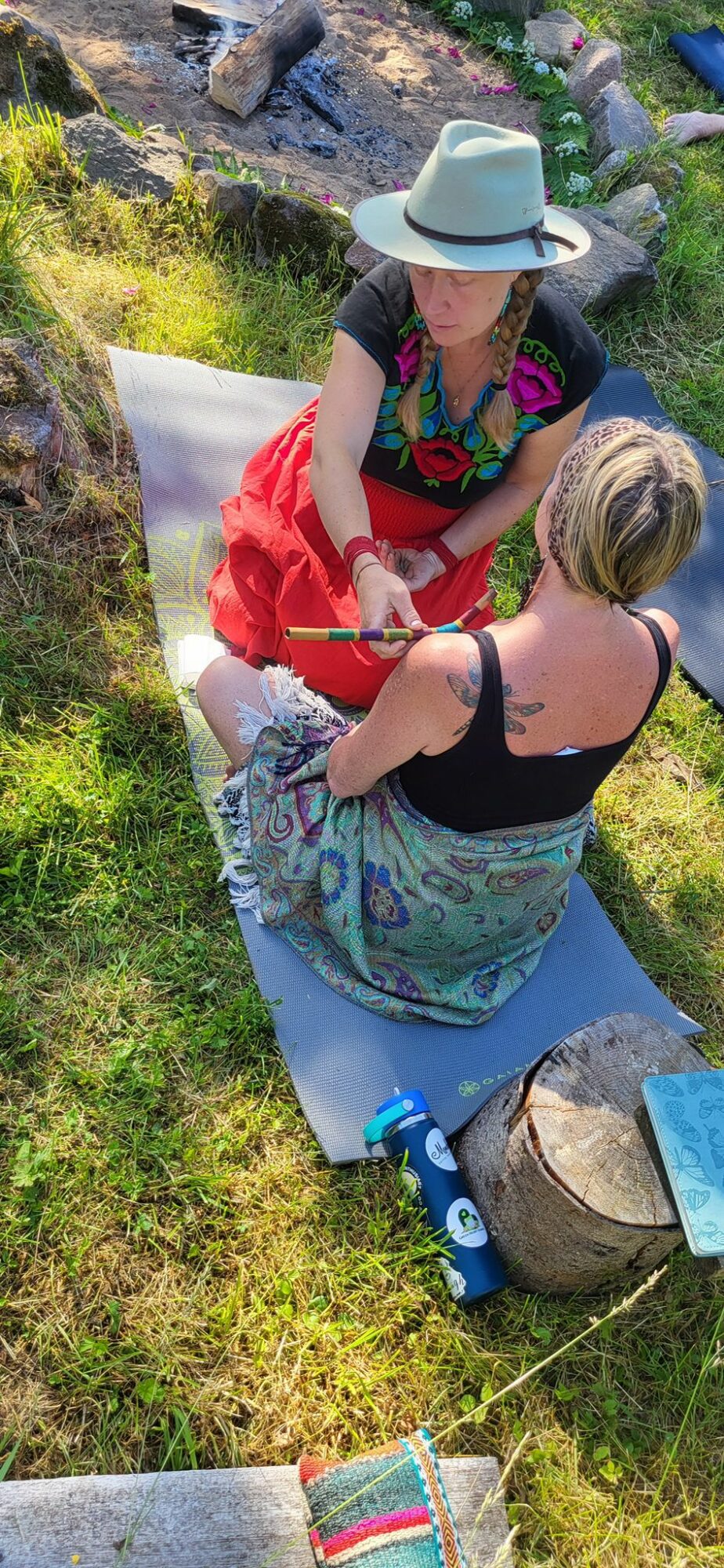 Two women sitting on a mat outdoors, one wearing a hat and colorful dress, the other in a black top and patterned skirt, talking.