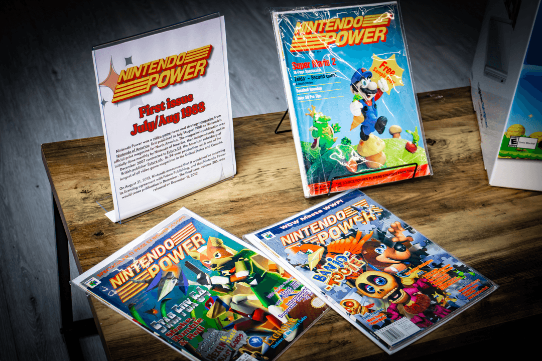 Four Nintendo Power magazines and a printed page on a wooden surface, with one magazine showing a character jumping over a river.