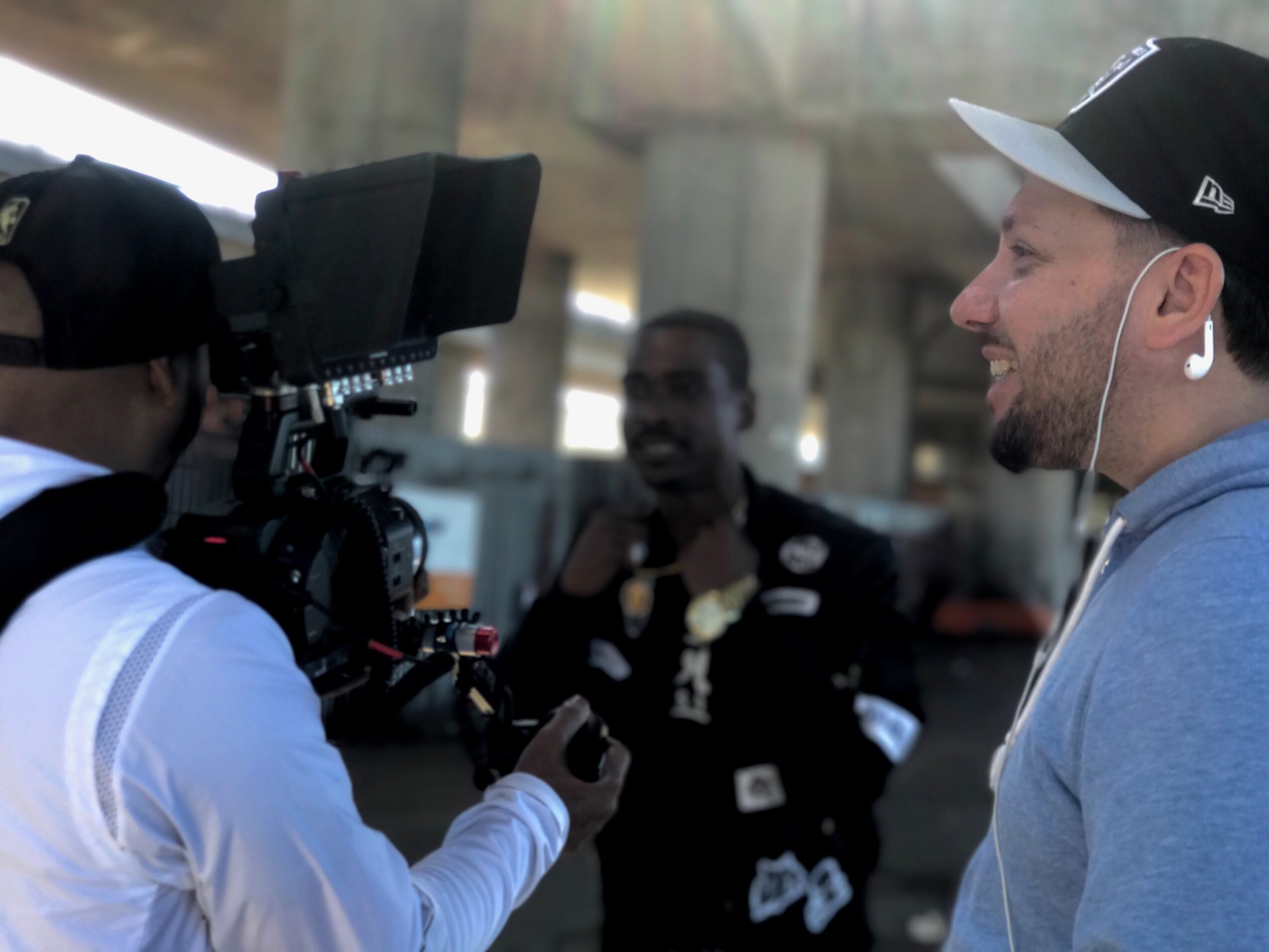 Two men filming a person under a bridge, one with a camera and the other with earphones, in an outdoor setting.