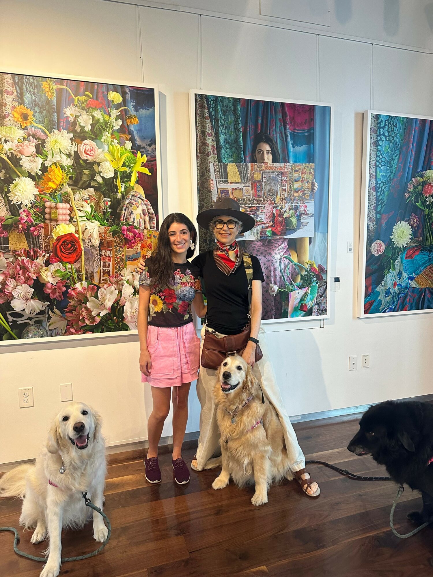 Two women and two dogs in an art gallery with colorful paintings and flowers, standing on wooden floor.