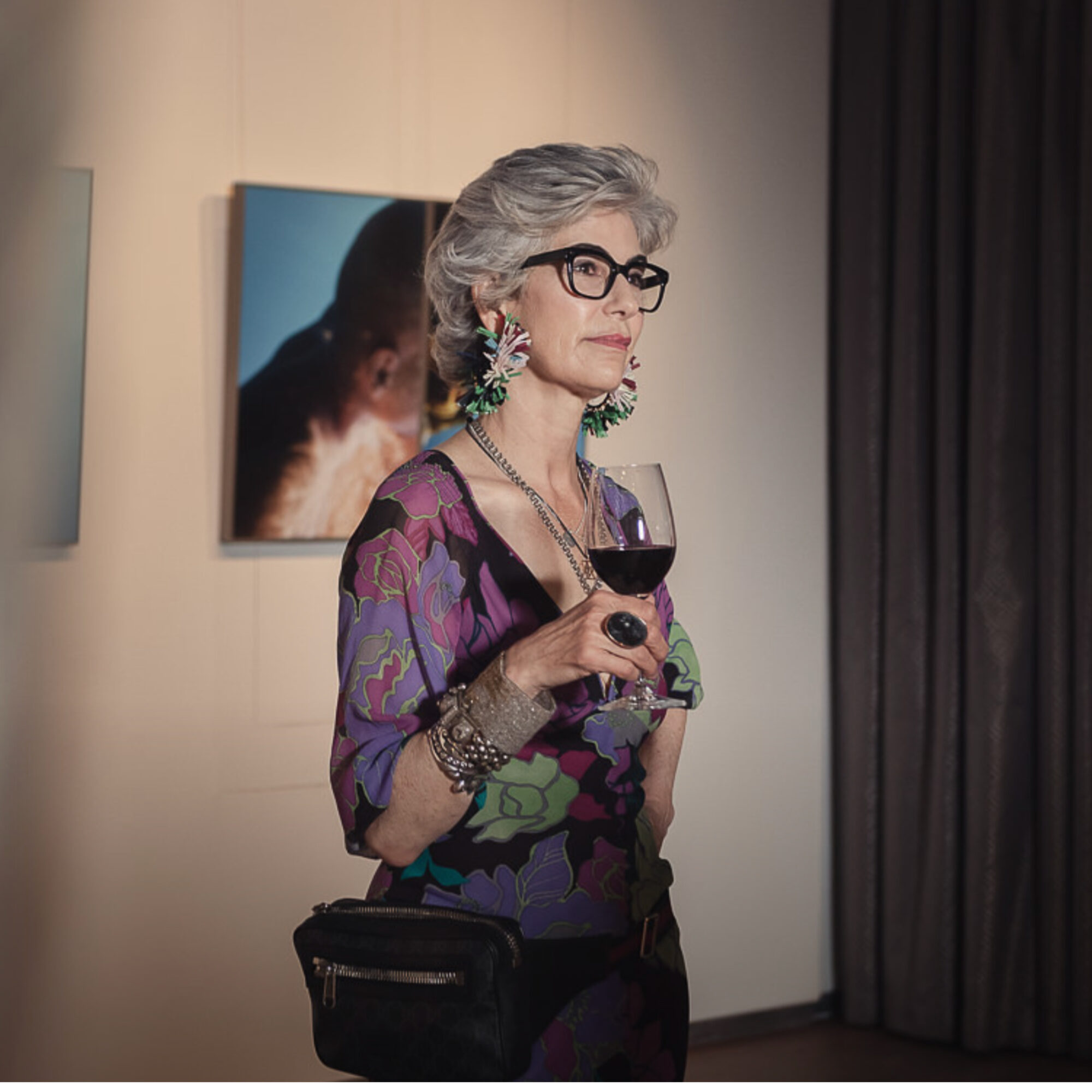 Older woman with gray hair, glasses, and earrings, holding a glass of red wine, standing indoors near a curtain.