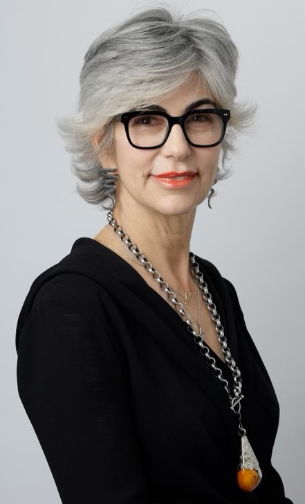 A woman with short gray hair, glasses, wearing a black top and a necklace, smiling slightly.
