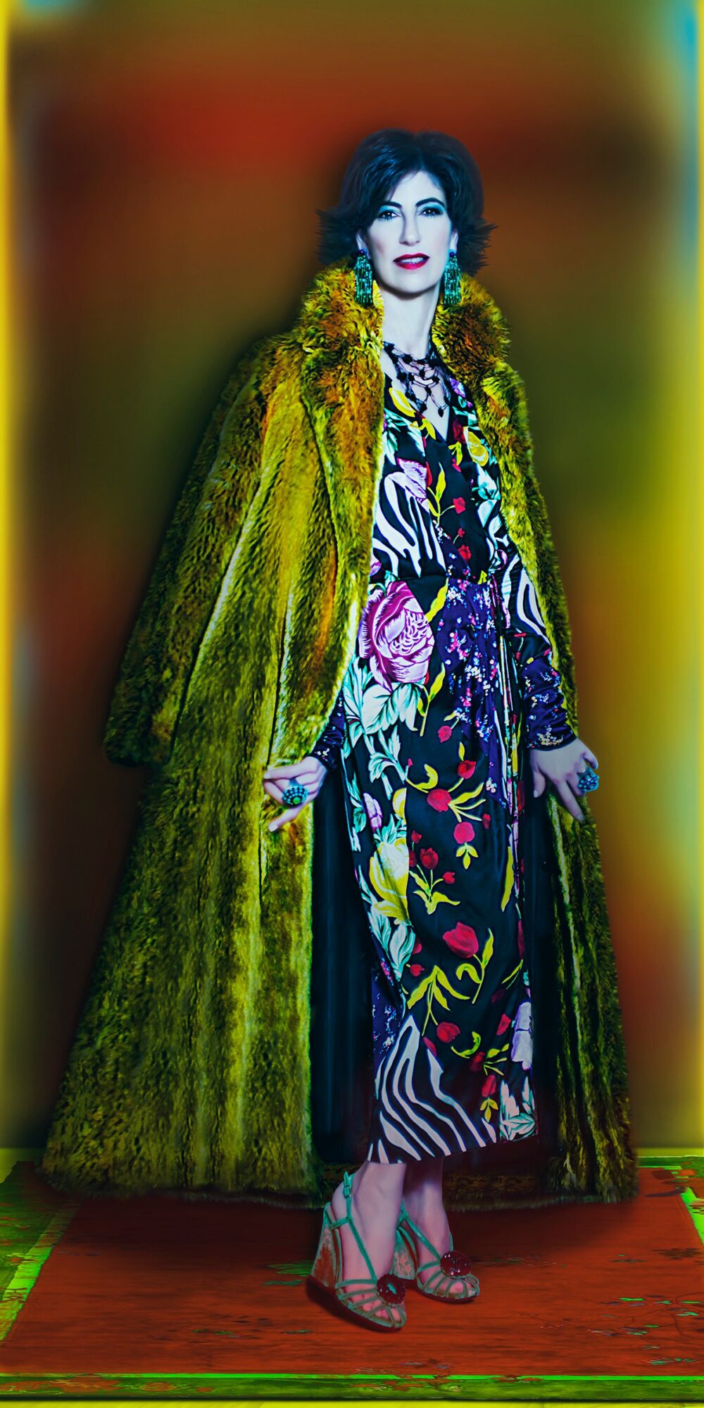 Woman in colorful patterned dress and yellow fur coat stands on a red carpet, with a blurred colorful background.