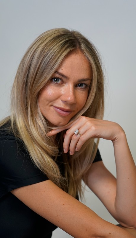 Woman with long blonde hair resting her chin on her hand, smiling softly, wearing a black top, against a plain background.