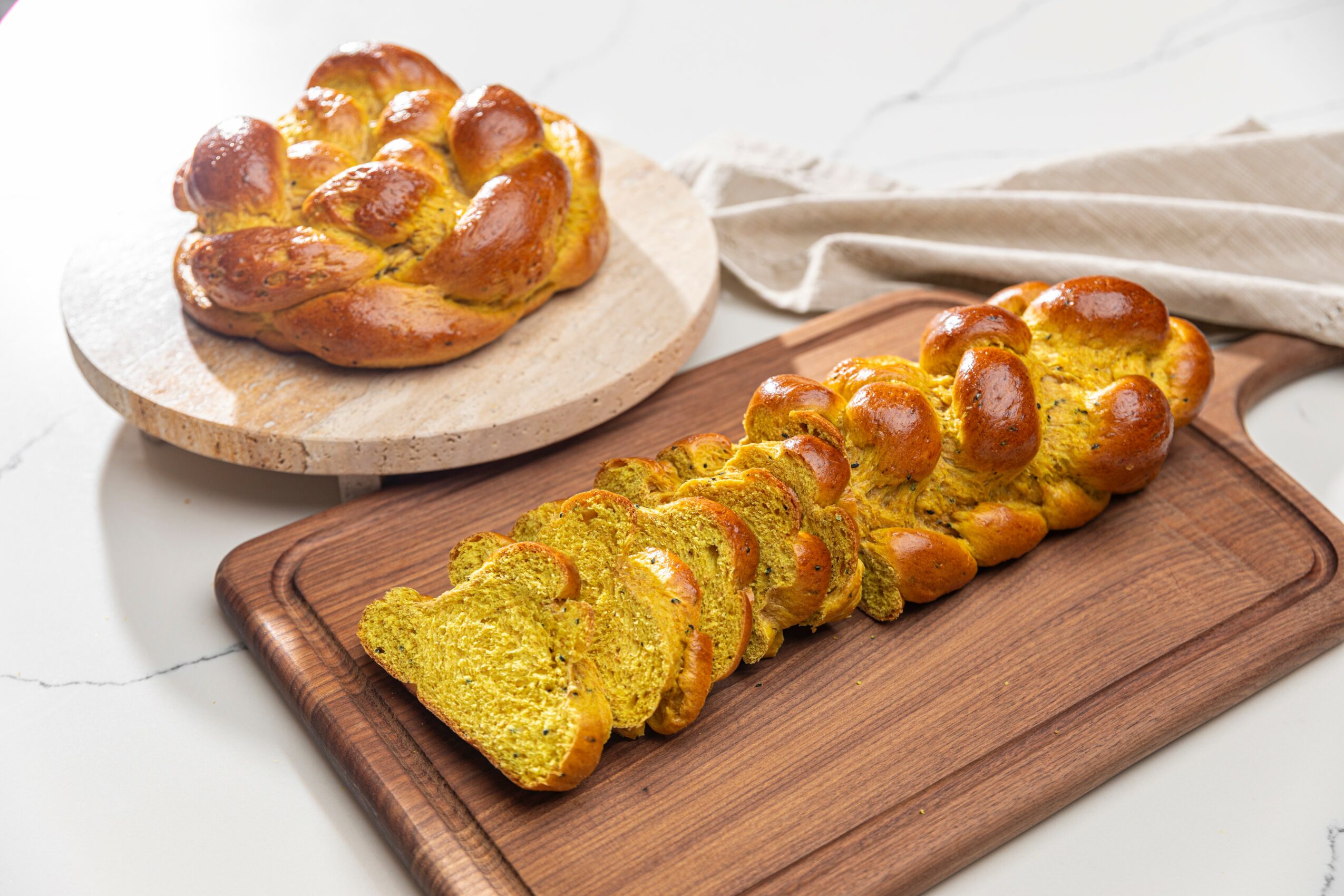 Loaf of sliced bread with a golden crust on a wooden cutting board, with a whole loaf in the background, on a white surface