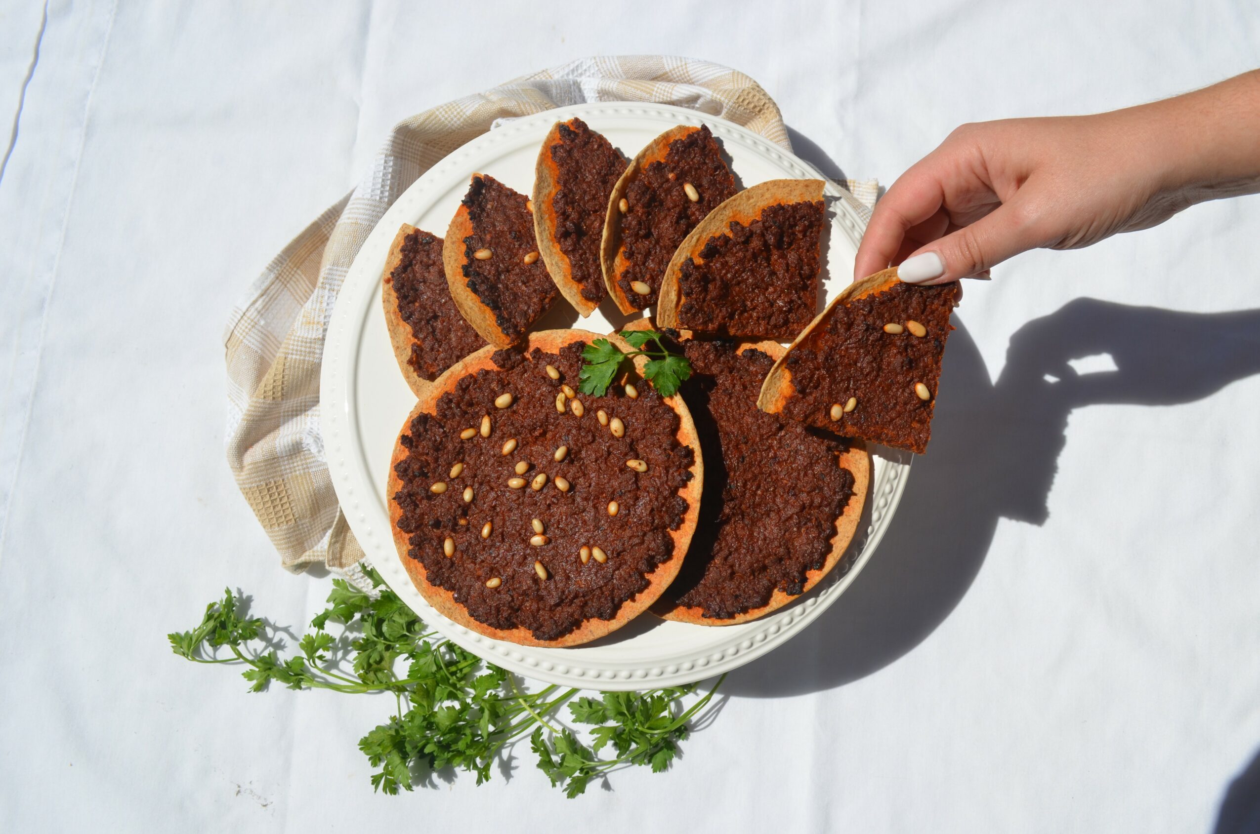 Hand reaching for a slice of round, dark-colored cake with nuts on a white plate, with green garnish, on a white surface.