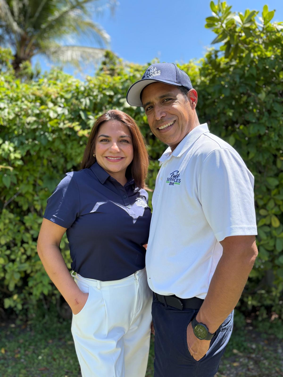 Two people standing outdoors with greenery and blue sky, smiling, one wearing a navy shirt and the other a white shirt and cap.