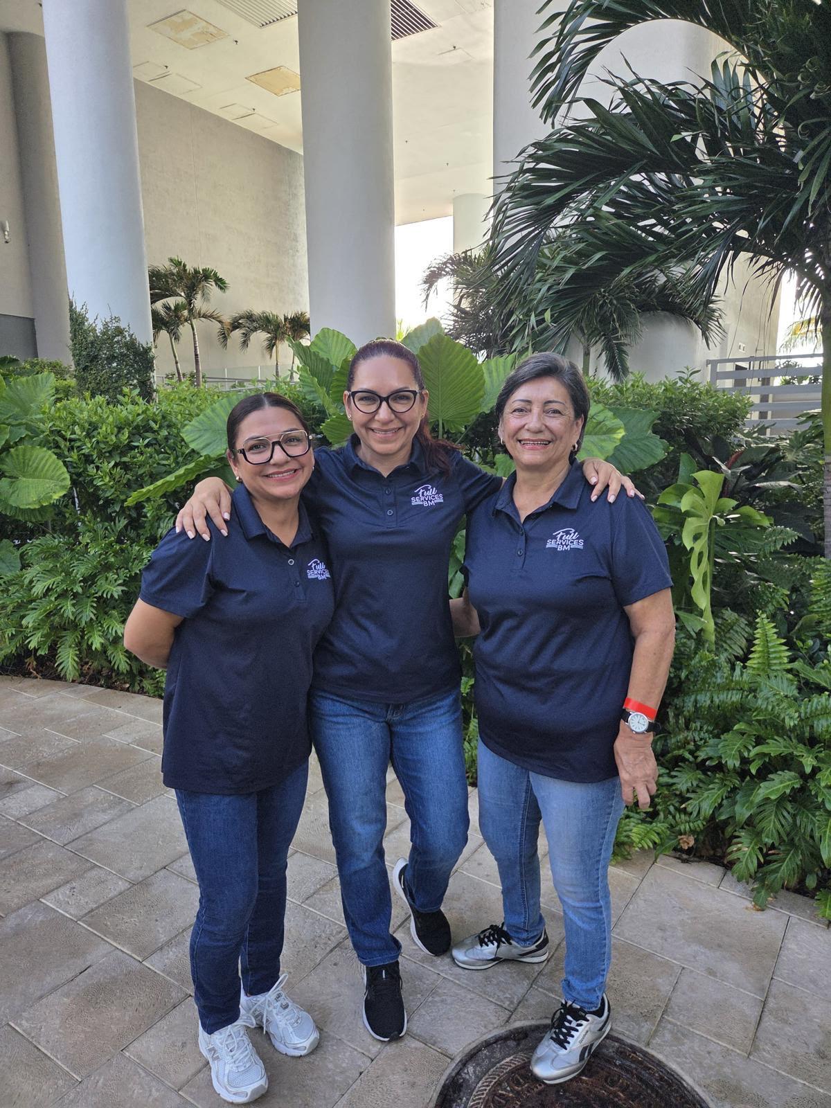 Three women in matching navy blue shirts and jeans standing outdoors with greenery and large columns behind them, smiling and embracing.