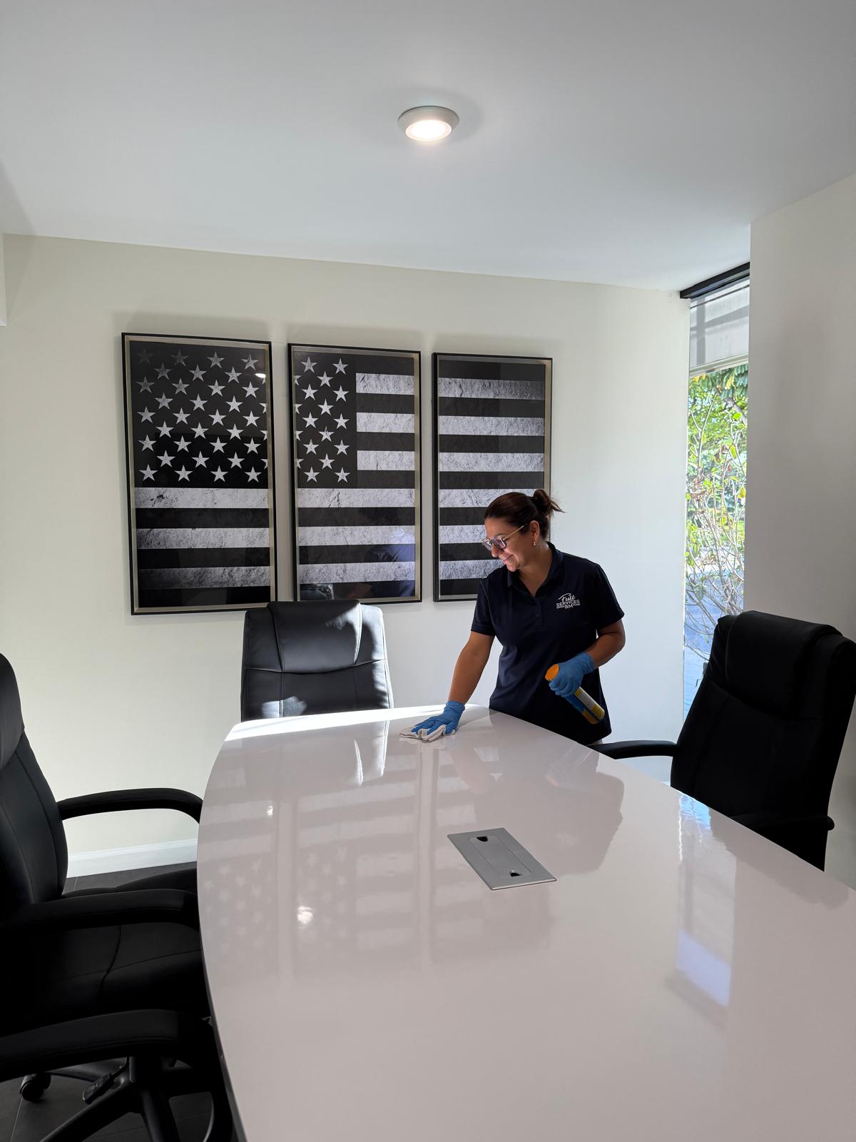 Woman cleaning a white conference table in a modern office with American flag artwork on the wall.