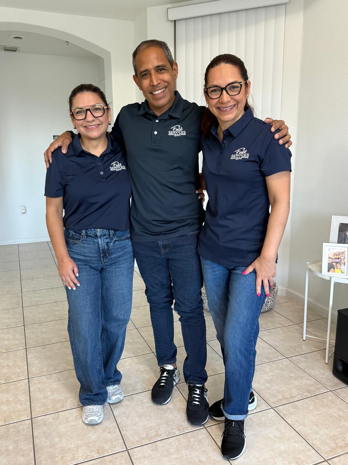 Three people standing indoors, smiling, with arms around each other, wearing matching navy blue shirts and jeans.