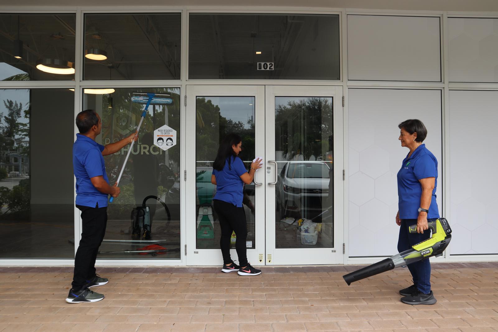 Three people standing outside a glass door, one holding a leaf blower, another using a spray bottle, and a third person in the middle.