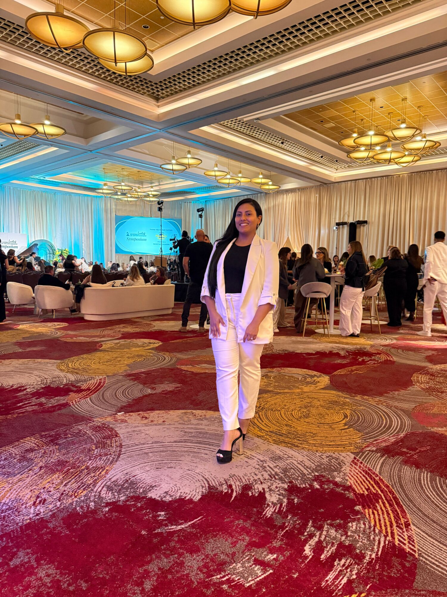 Woman in white pants and blazer stands on a patterned carpet at an indoor event, with people and tables in background.